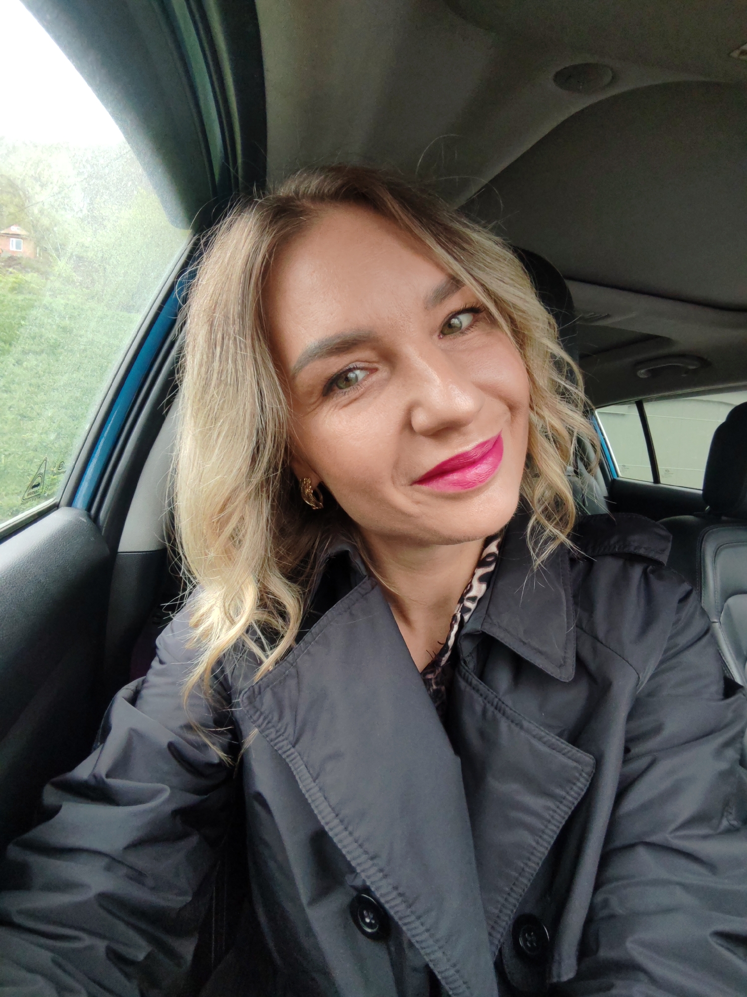 A blonde woman with wavy hair, green eyes, and bright pink lipstick smiles cheerfully while posing for a selfie inside a car. She is seated in what appears to be the front passenger seat, wearing a black trench coat over a leopard print top. A gold-toned, twisted earring is visible on her right ear.
The car's interior, including the headliner, seat, and window frame, is visible. Through the car window, a lush green outdoor environment can be seen, featuring what looks like hills or trees. Faint streaks on the window suggest either rain or condensation. A small, light-colored building with dark windows is partially visible in the distant background through the greenery. The lighting indicates it is daytime, likely overcast. Faint, unreadable markings, possibly from a car sticker, are present on the window glass. The woman's expression conveys happiness or contentment.