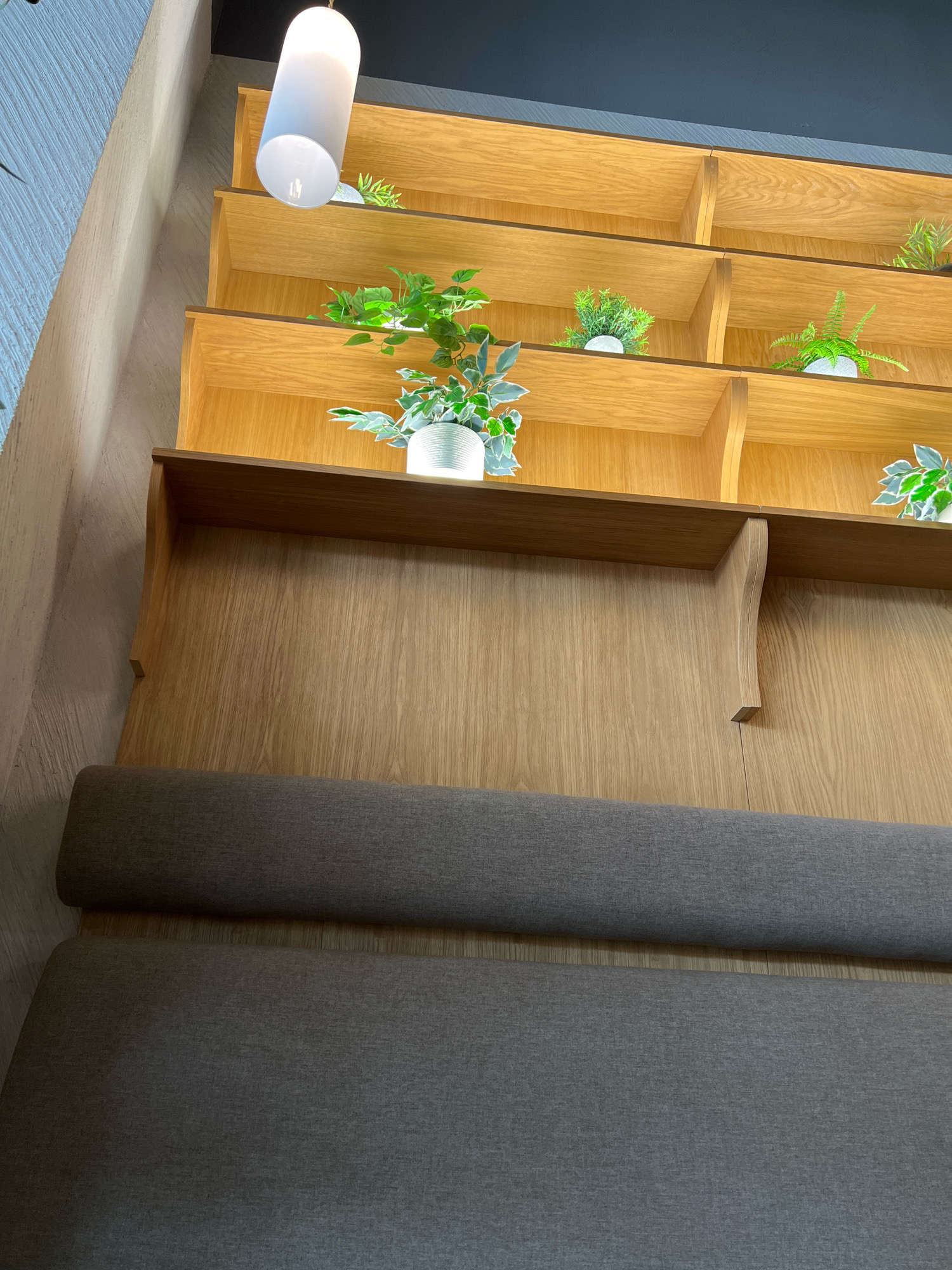 This is a close-up, low-angle shot of a modern interior featuring wooden shelves filled with small potted plants. The shelves are illuminated from above by a cylindrical pendant light. Below the shelves, a dark gray upholstered bench or sofa is visible. The overall aesthetic suggests a contemporary and minimalist design. The image does not contain any information to determine its specific location within Krasnoyarsk, Russia.