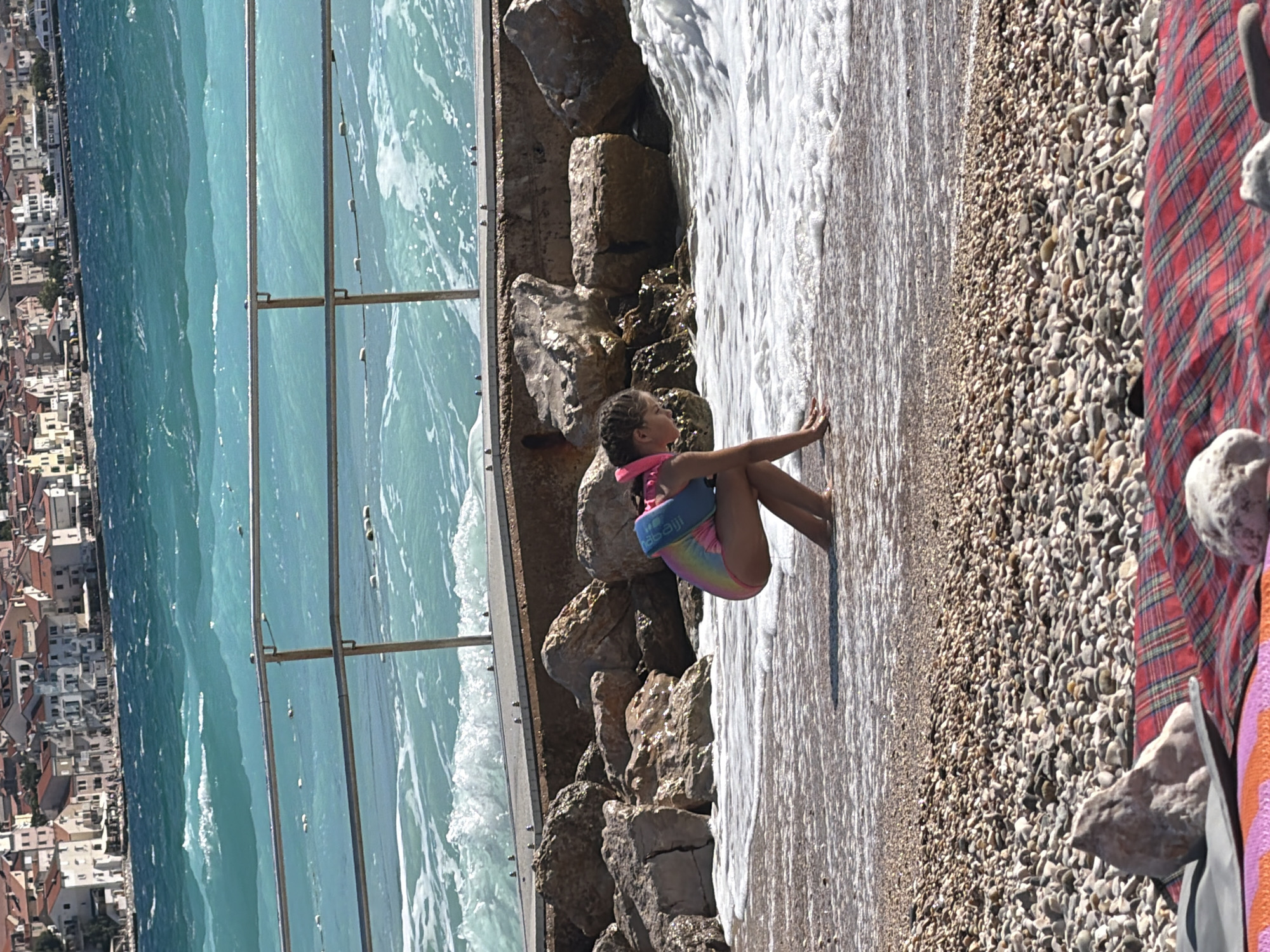 A young girl sits on a pebble beach, her legs extended towards the water. She wears a colorful swimsuit and a buoyancy aid around her torso, which appears to be a light blue float with some text that may be a brand name. Waves gently lap at the shore, creating a foamy white edge that reaches her feet. To her left, a rocky embankment rises, with larger stones forming a natural barrier. Beyond the embankment, a metal railing separates the beach from a body of water that is a vibrant turquoise color, reflecting the clear sky. In the distance, a town with white and terracotta-roofed buildings climbs up a hillside. The scene is bathed in bright sunlight, suggesting a warm, sunny day, likely midday. The girl appears relaxed, with her right arm outstretched towards the water. The overall impression is one of a peaceful summer day at the seaside.
