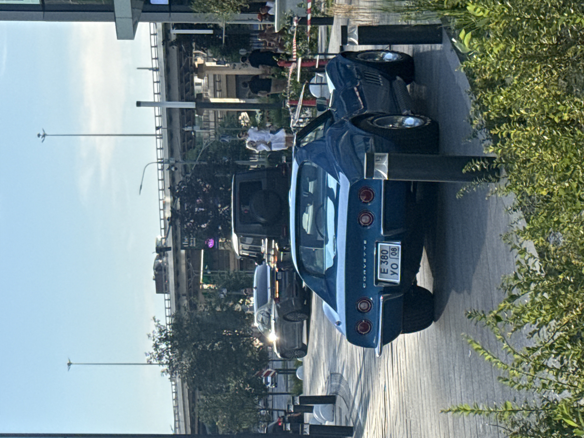 In the city of Khimki, Russia, a blue Chevrolet Corvette is parked on a paved area with decorative bollards. The car has a license plate reading "E 380 YO 08". Behind the Corvette, a silver sedan is visible. Further in the background, a dark SUV is parked near a group of people. A pedestrian bridge with streetlights and trees are prominent elements of the urban landscape. The scene is illuminated by natural sunlight under a clear blue sky.