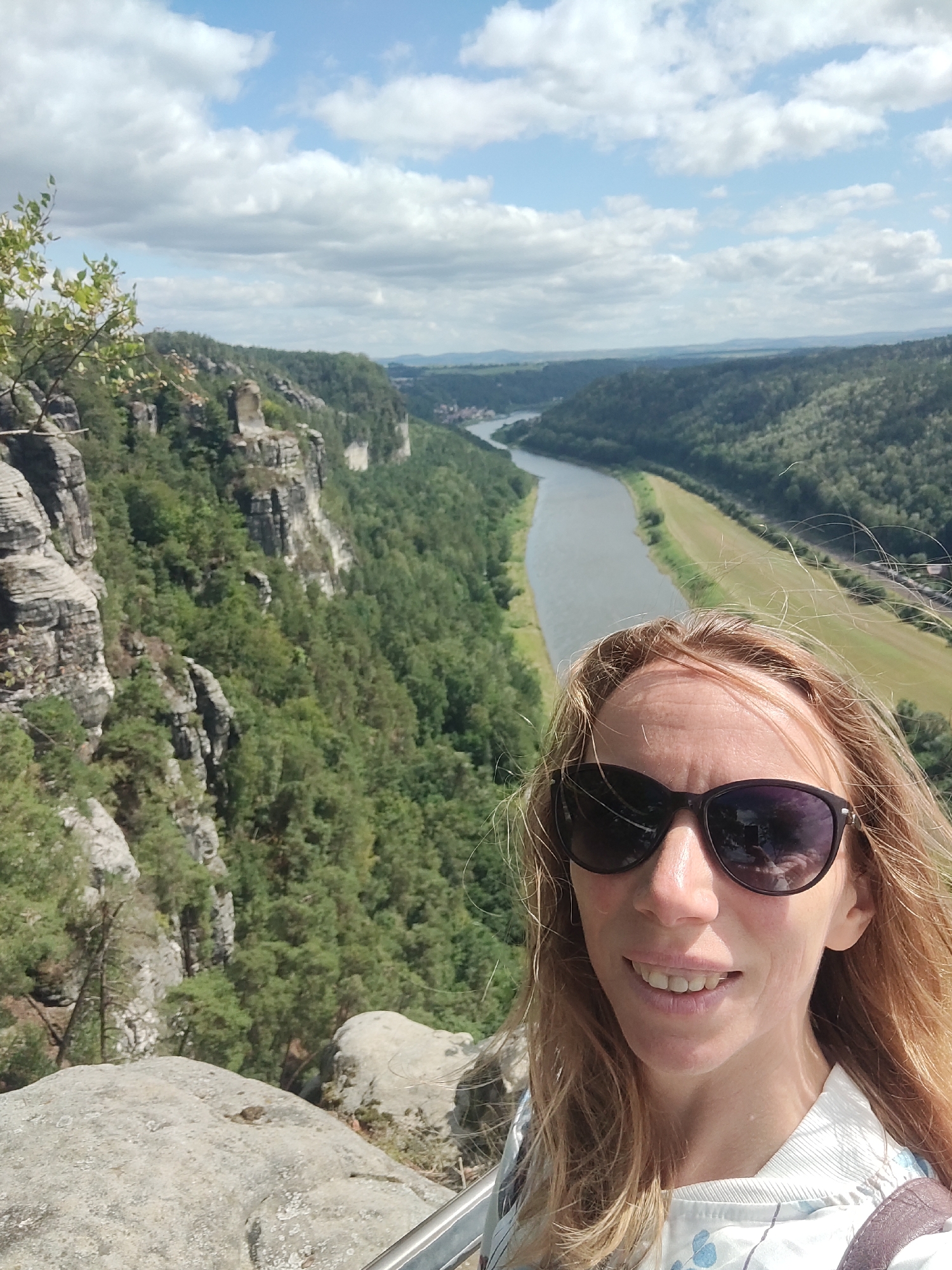 A selfie is taken by a blonde woman with light skin, wearing sunglasses and a white shirt with a floral pattern. She is smiling. The background is a scenic view of the Elbe River valley near Lohmen, Germany. The valley is characterized by steep, forested cliffs on the left and a wide, calm river with a grassy embankment on the right. The sky is partly cloudy. The woman is standing on a rocky outcrop.