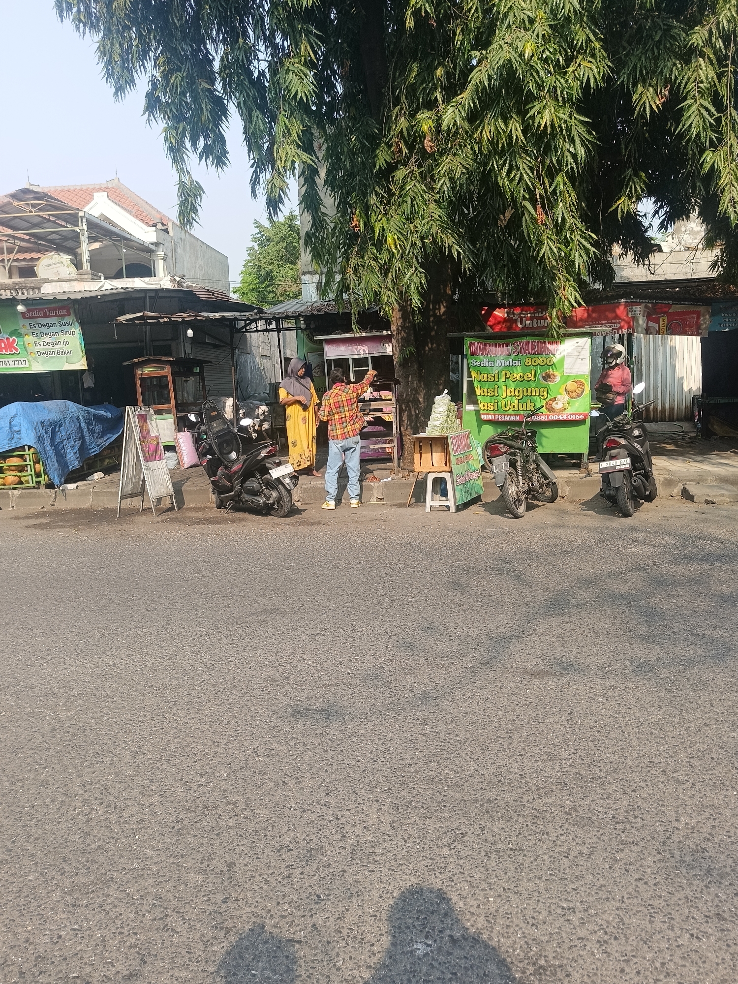 The image shows a street scene in Wedi, Indonesia, on a sunny day. A large tree with dense foliage dominates the upper part of the frame, casting shadows on the road and sidewalk. Several small food stalls are set up along the roadside, some with green banners advertising various food and drink options. One banner reads "WARUNG SYAKINAH" and lists "Nasi Pecel" (rice with peanut sauce salad) and "Nasi Uduk" (coconut rice) as offerings.
Two individuals are interacting near one of the stalls. A woman in a yellow hijab and dress is standing, and a man in a plaid shirt and jeans is facing away from the camera, pointing towards something on the stall. Two motorcycles are parked on the sidewalk. One is a black scooter parked upright, and another black motorcycle is parked on its side. In the background, there are buildings with red-tiled roofs and more stalls or shops. A blue tarp covers what appears to be stacked produce or goods on a cart. The road in the foreground is asphalt, with shadows of people and objects visible on its surface.