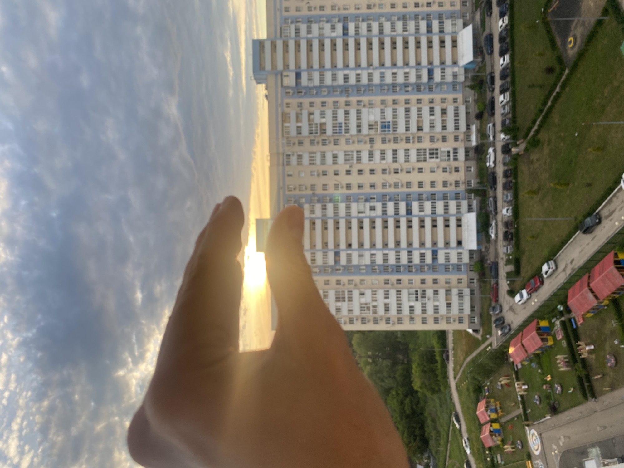 This media file is an aerial, upside-down shot of a residential area in Novokuybyshevsk, Russia, featuring a large, multi-story apartment building. In the foreground, a person's hands are obscuring part of the view, with one hand appearing to be held up towards the sky, possibly framing the scene or blocking the sun. The sky is cloudy, with the sun casting a bright glow, suggesting it is either sunrise or sunset.
The apartment building is light-colored with blue accents and numerous windows, indicating a densely populated urban environment. Below and to the right of the building, a green grassy area is visible, dotted with trees and several children's playgrounds, characterized by colorful structures with red roofs. A paved road runs alongside the grassy area, with a line of parked cars visible. The overall scene depicts a typical urban residential neighborhood.
The time of day is likely early morning or late evening due to the low angle of the sun and its bright, diffused light. The weather appears clear apart from the clouds. No visible text is present in the image. The emotion conveyed is one of observation and perhaps a sense of scale, with the human hands providing a point of reference against the vastness of the building and sky.