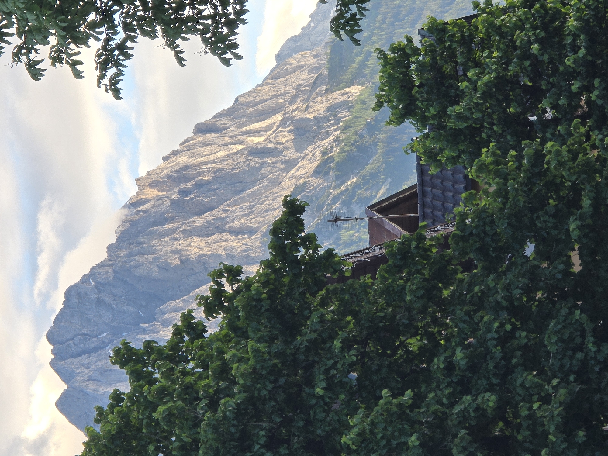 This image depicts a mountainous landscape in the vicinity of Garmisch-Partenkirchen, Germany, framed by dense green foliage. In the background, prominent, rugged grey mountains with distinct rocky ridges and some lighter rock or snow patches dominate the upper and mid-sections of the frame. Sunlight illuminates the mountain faces, highlighting their texture and creating contrasts.
In the mid-ground, a substantial portion of the scene is filled with the vibrant green leaves of deciduous trees, partially obscuring other elements. Through these trees, in the lower-mid section of the image, the dark tiled roof of a building is visible. A small spire-like structure topped with a dark cross rises from the roofline towards the center. To the left of this cross, a multi-pronged antenna is mounted on the roof. The building's architecture, combined with the visible cross, suggests it may be a local chapel or a traditional Alpine house.
The sky above the mountains is partly cloudy, featuring patches of light blue sky interspersed with white and grey clouds. The overall lighting suggests either early morning or late afternoon. No human figures or discernible actions are visible. The scene conveys a serene, natural environment typical of the Bavarian Alps around Garmisch-Partenkirchen.