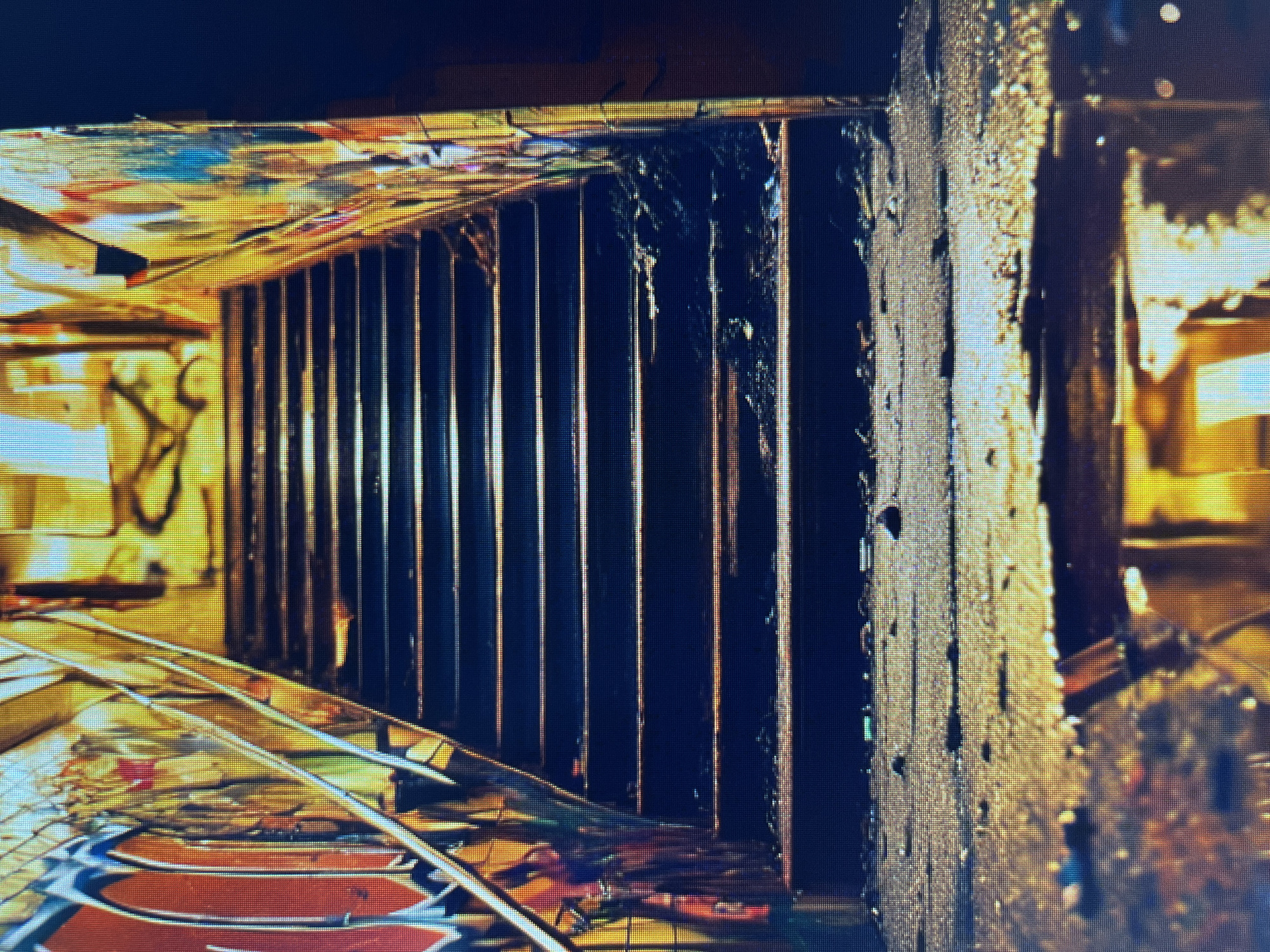 The image depicts a set of dark, weathered stairs leading upwards, flanked by graffiti-covered walls. The stairs appear to be made of concrete or stone and are littered with debris, including leaves and dirt. A metal handrail runs along the left side of the staircase. The walls on either side are extensively covered in vibrant and colorful graffiti art. The lighting in the scene is dramatic, with strong yellow and orange hues casting long shadows, suggesting either artificial lighting or the setting sun. At the bottom of the stairs, the ground is wet and reflective, with puddles of water mirroring the surrounding light and structures. The overall impression is one of urban decay and artistic expression in a gritty, outdoor environment.
