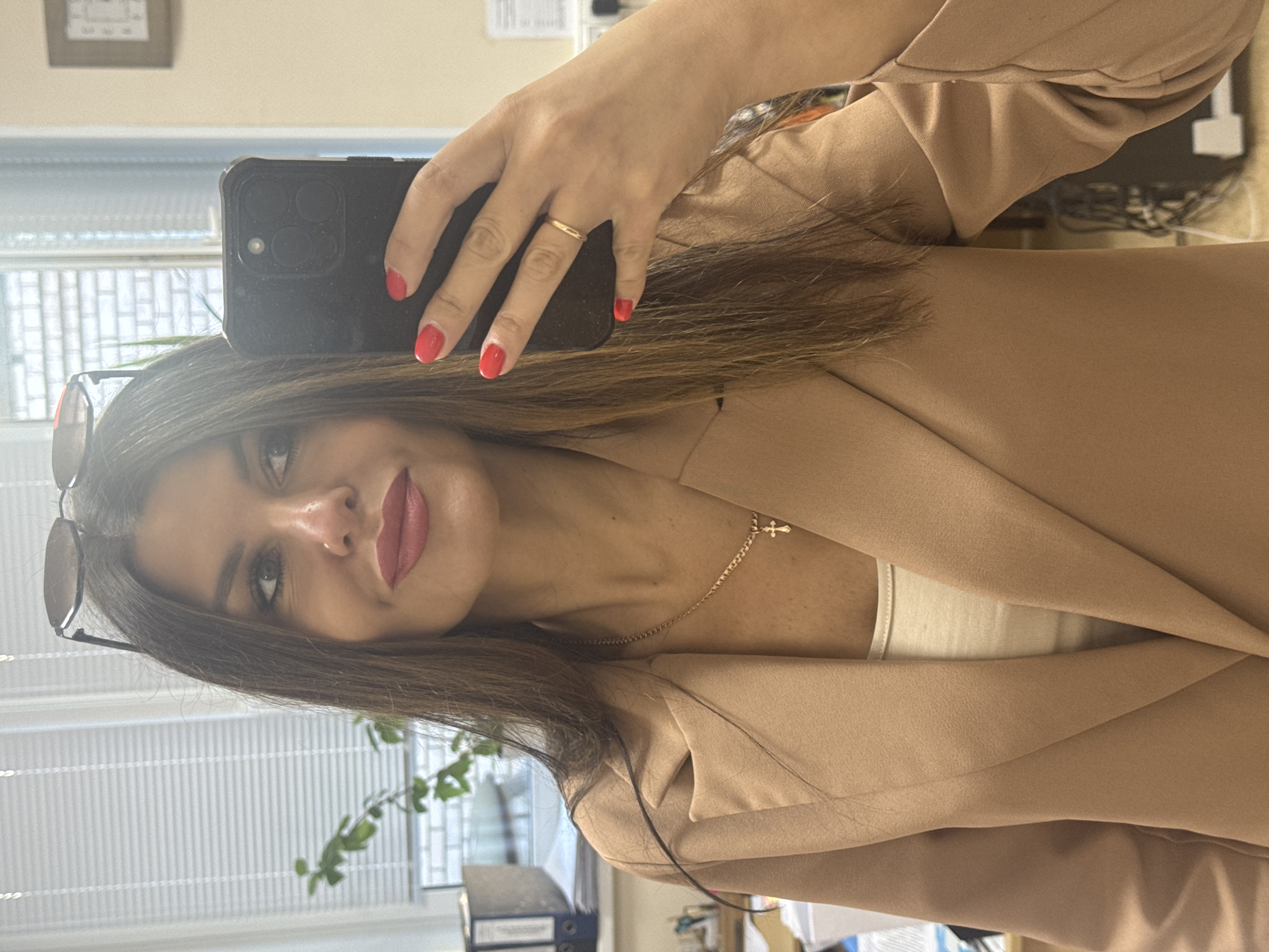 A woman is taking a selfie in an office environment. She has long, dark brown hair, and is wearing red lipstick and nail polish. She is wearing a beige blazer over a cream-colored top, and a gold chain with a cross pendant. She is also wearing a thin gold ring on her left hand. She is holding a black smartphone in her right hand to take the photo. Behind her, a window with blinds and a plant are visible. A filing cabinet is also visible in the background. The overall impression is that of a professional setting. The location is not specified as Chaikovsky, Russia.