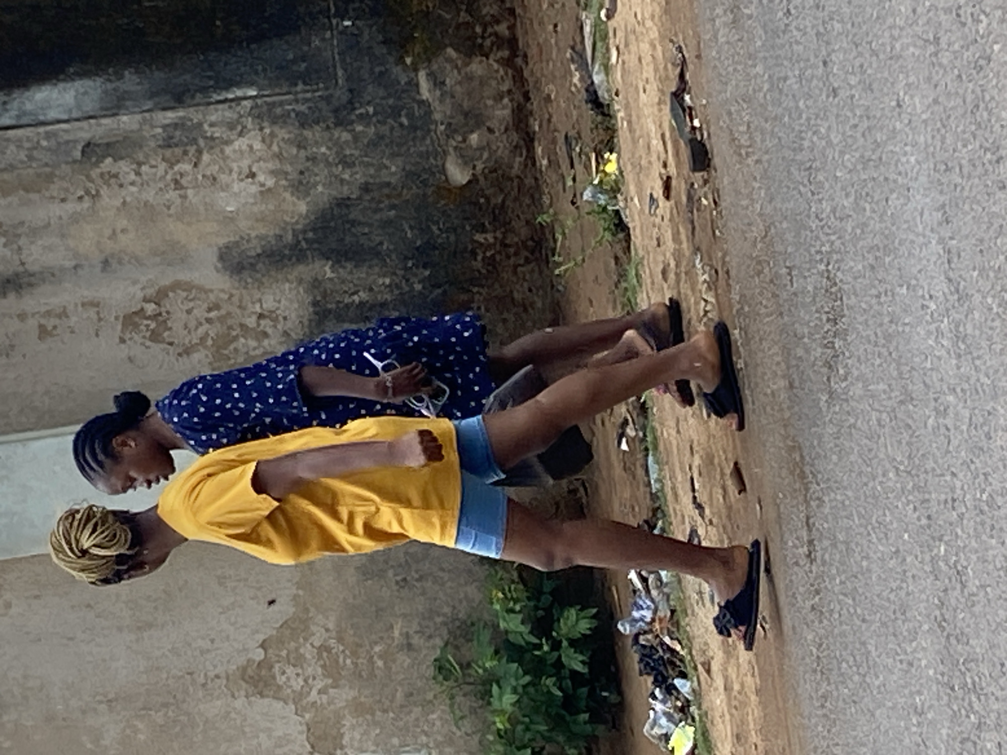 Two individuals walk on an asphalt road alongside a dilapidated concrete structure. The person in the foreground wears a yellow shirt and blue shorts, while the other is in a blue patterned dress. Both wear dark-colored sandals. Vegetation and scattered trash are present on the roadside. The image is from Narayi, Nigeria.