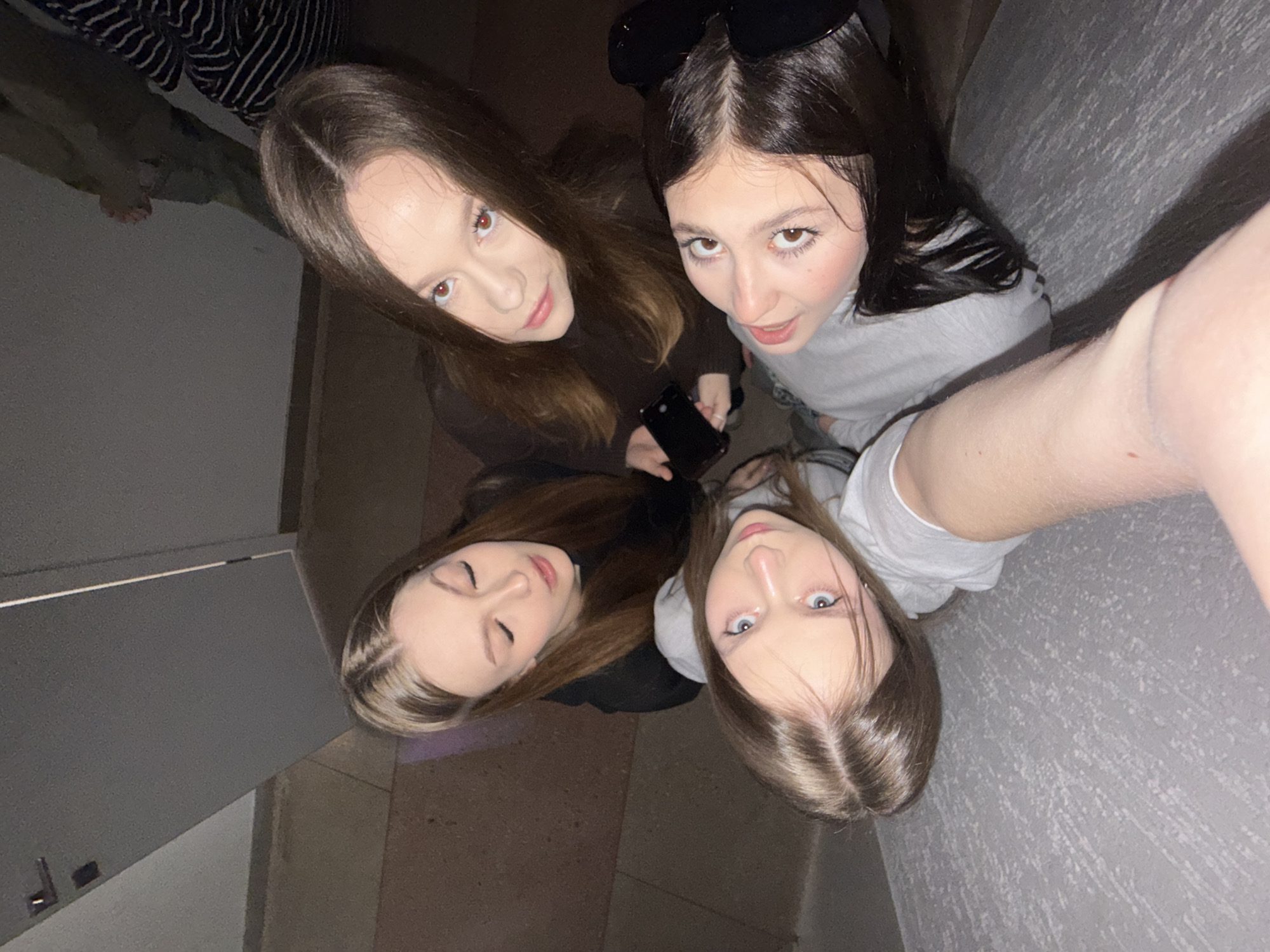 A high-angle shot depicts four young women. One of the women is holding a black phone, likely taking the picture. The location appears to be indoors, indicated by the type of flooring and wall textures. The image was presumably taken in the city of Chaikovsky, Russia. One can see an edge of the door and striped clothes in the left side of the picture.