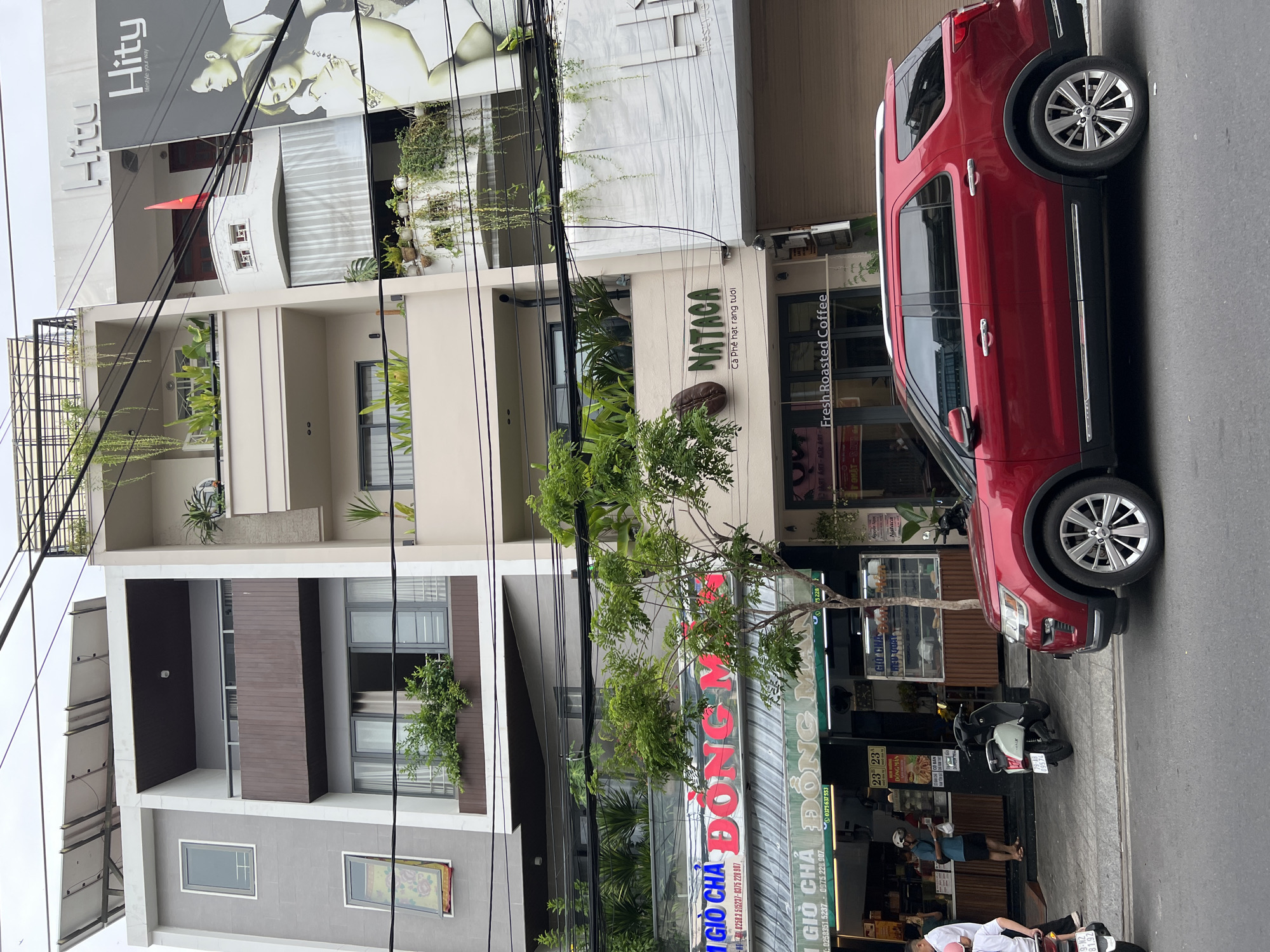 In Nha Trang, Vietnam, a red SUV is parked on the street in front of a row of buildings. The buildings appear to be shops or cafes, with signage in Vietnamese and English. One sign reads "DONG MAN" in large red letters, another says "NATACA Fresh Roasted Coffee." People are visible in the doorways of the shops, and motorbikes are parked along the sidewalk. Lush green plants cascade from balconies and hang from the buildings, suggesting a tropical urban environment. Power lines crisscross the sky above the street. The buildings have modern architecture, with balconies and large windows. The weather appears to be overcast.