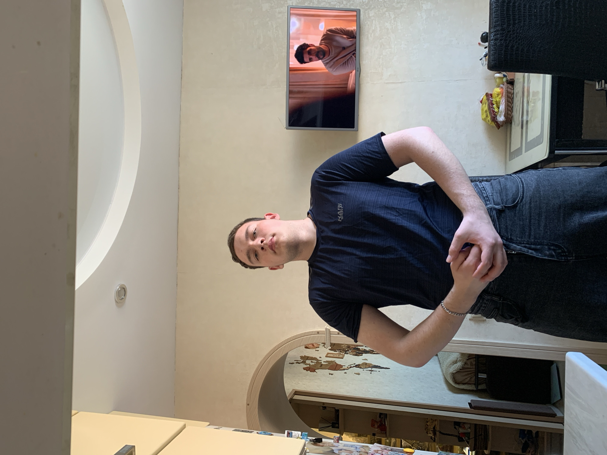 A young man stands indoors, looking directly at the camera with a neutral expression. He is wearing a dark blue T-shirt with the text "HARI SPORT" visible on his chest, and dark jeans. His hands are clasped together in front of his waist.
The background shows a room that appears to be a living area or office in Severodvinsk, Russia. To the man's left, a flat-screen television is mounted on a textured wall, displaying a video of another man with a beard, wearing a light-colored top. To the man's right, an arched doorway leads into an adjacent room, where a decorative wooden world map is affixed to the wall. Above the man's head, there is a distinct curved architectural feature on the ceiling or upper wall, and a circular light fixture.
Various objects are partially visible around the room. On the right, part of a dark, possibly leather-textured piece of furniture (like a chair or sofa) is seen, with a basket containing items underneath it. In the foreground, a light-colored table or counter surface is present. The walls have a light, textured finish. The room is brightly lit, suggesting daytime or good interior lighting.