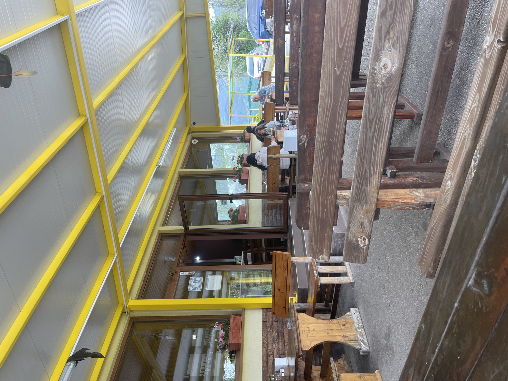 The image shows the outdoor seating area of a restaurant, likely in Sheynovo, Bulgaria, given the prompt's request for location information. The seating area is covered by a metal awning with a white roof supported by yellow beams. Several wooden tables and benches are visible. The foreground tables are arranged vertically in the image. People are seated at some of the tables in the background. The restaurant has glass windows and doors. Flower boxes are placed beneath some windows. Beyond the restaurant, trees and vehicles are visible.