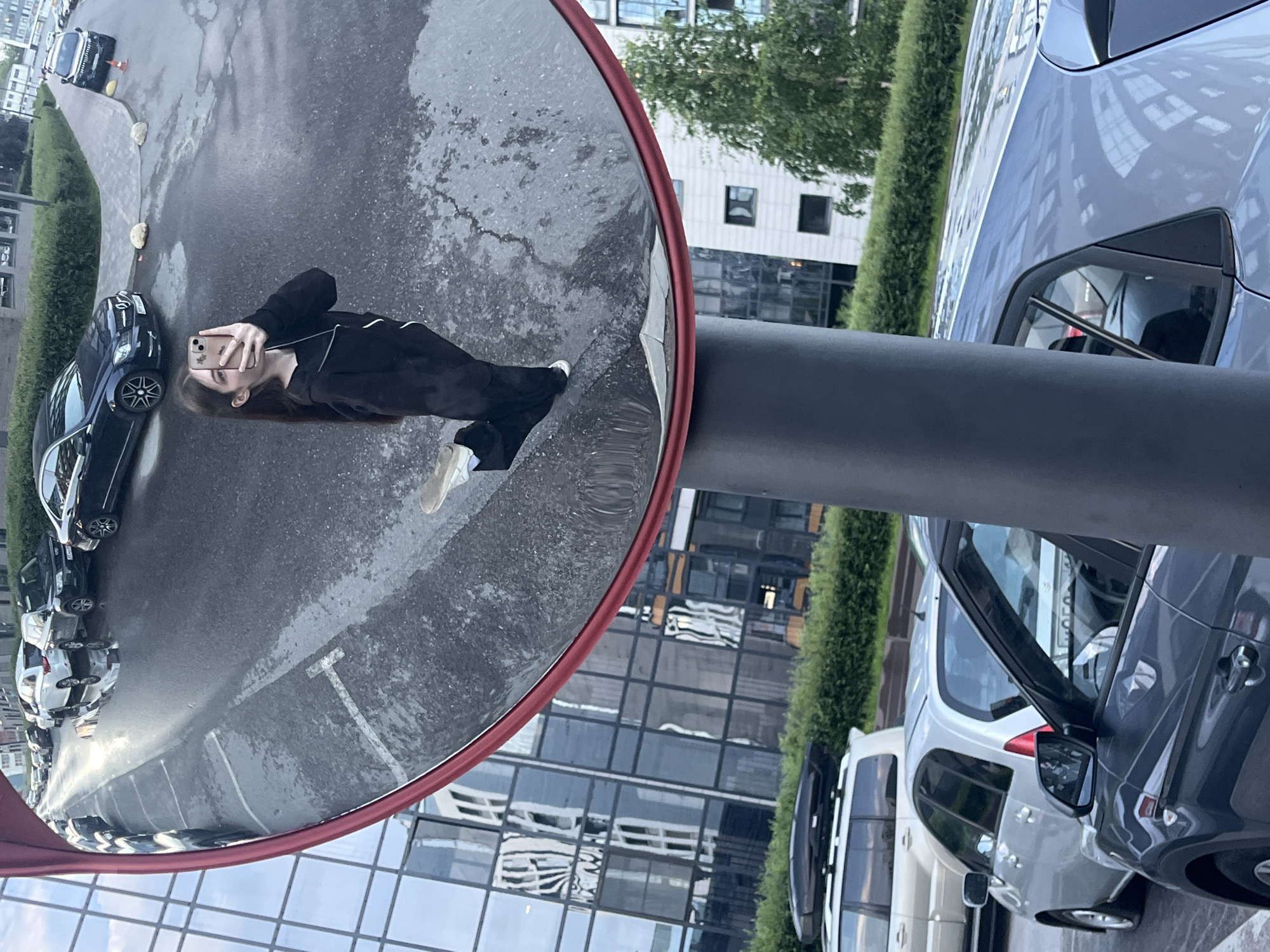 A full-length, distorted selfie is taken by a young woman using her smartphone, reflected in a large, convex security mirror. She is wearing a black hooded sweatshirt and black pants with white accents, and white sneakers. The mirror is mounted on a dark gray pole and has a red trim. In the background, reflected in the mirror, are several cars parked on a wet asphalt street, including a dark blue sedan, a white SUV, and other vehicles. The reflection also shows a sidewalk, green bushes, and the facades of modern buildings with large glass windows. In the foreground, outside the mirror's reflection, parts of several cars are visible, including a silver hatchback and a dark gray car. The setting appears to be an urban environment.