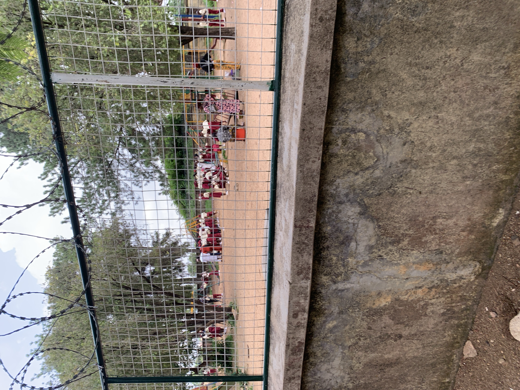 The image shows a fenced-in school playground area, possibly in Jos, Nigeria. The fence in the foreground is a metal mesh with barbed wire on top. Beyond the fence, children in school uniforms, primarily red and white, are visible on a sandy playground. Some children are gathered in groups, while others appear to be interacting with playground equipment, such as a slide or a seesaw. Adults are also present, some observing the children and others engaged in conversation. The playground is surrounded by trees and vegetation. The ground appears to be made of sand, and there are some playground structures. The sky is overcast. The overall scene depicts a typical school day in an outdoor setting.