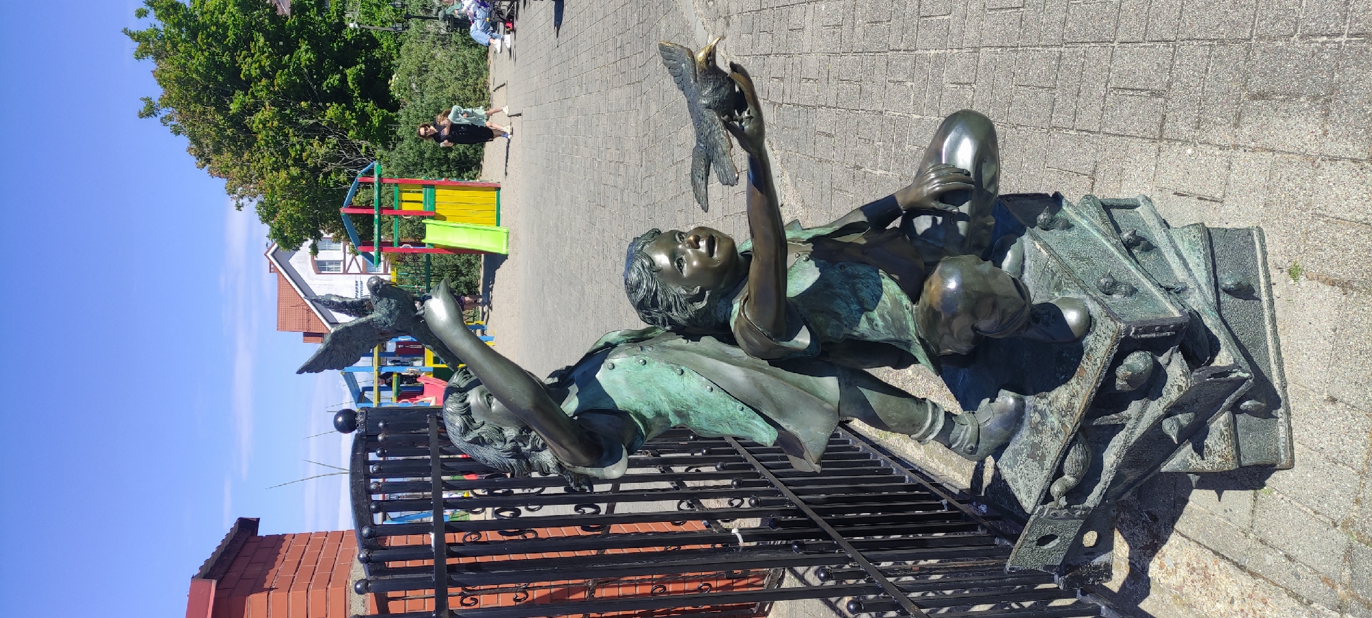 The image shows a bronze sculpture of two children interacting with birds. The taller child, on the left, is standing and reaching out with one arm towards a bird in flight. The shorter child, on the right, is kneeling and holding a bird in their outstretched hand. Both children are depicted with curly hair and wearing clothing that appears to be period attire. The sculpture is mounted on a base that resembles stacked, open crates, with smaller bird figures integrated into its design.
In the background, a playground with colorful slides and structures is visible. A woman in a black dress and a young girl in a light-colored dress are walking in the vicinity of the playground. Further in the background, there are buildings with red-tiled roofs and trees. The scene is set outdoors on a paved walkway under a clear blue sky.
This sculpture is located in the city of Zelenogradsk, Russia, on the promenade near the Baltic Sea.