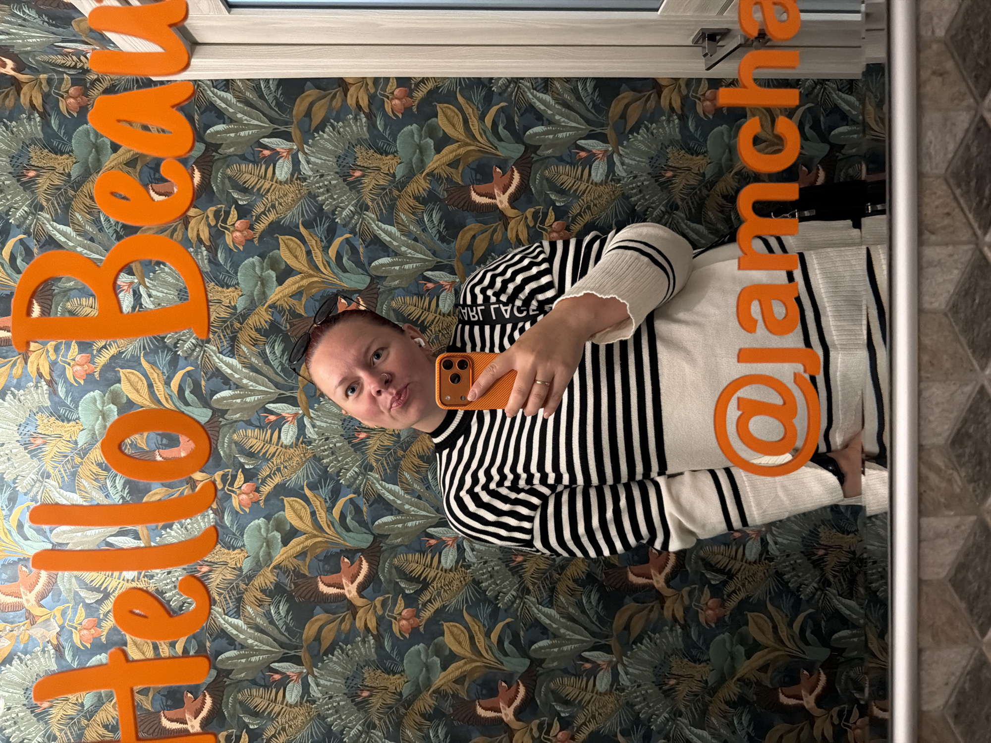 A mirror selfie features a woman holding an orange smartphone. She has fair skin, reddish hair, sunglasses perched on her head, and a nose ring. She wears a black and white striped sweater. On the mirror, orange lettering spells out "Hello Bear" and "@iamchka." The background features patterned wallpaper with leaves and bird motifs. The location is Chaikovsky, Russia.