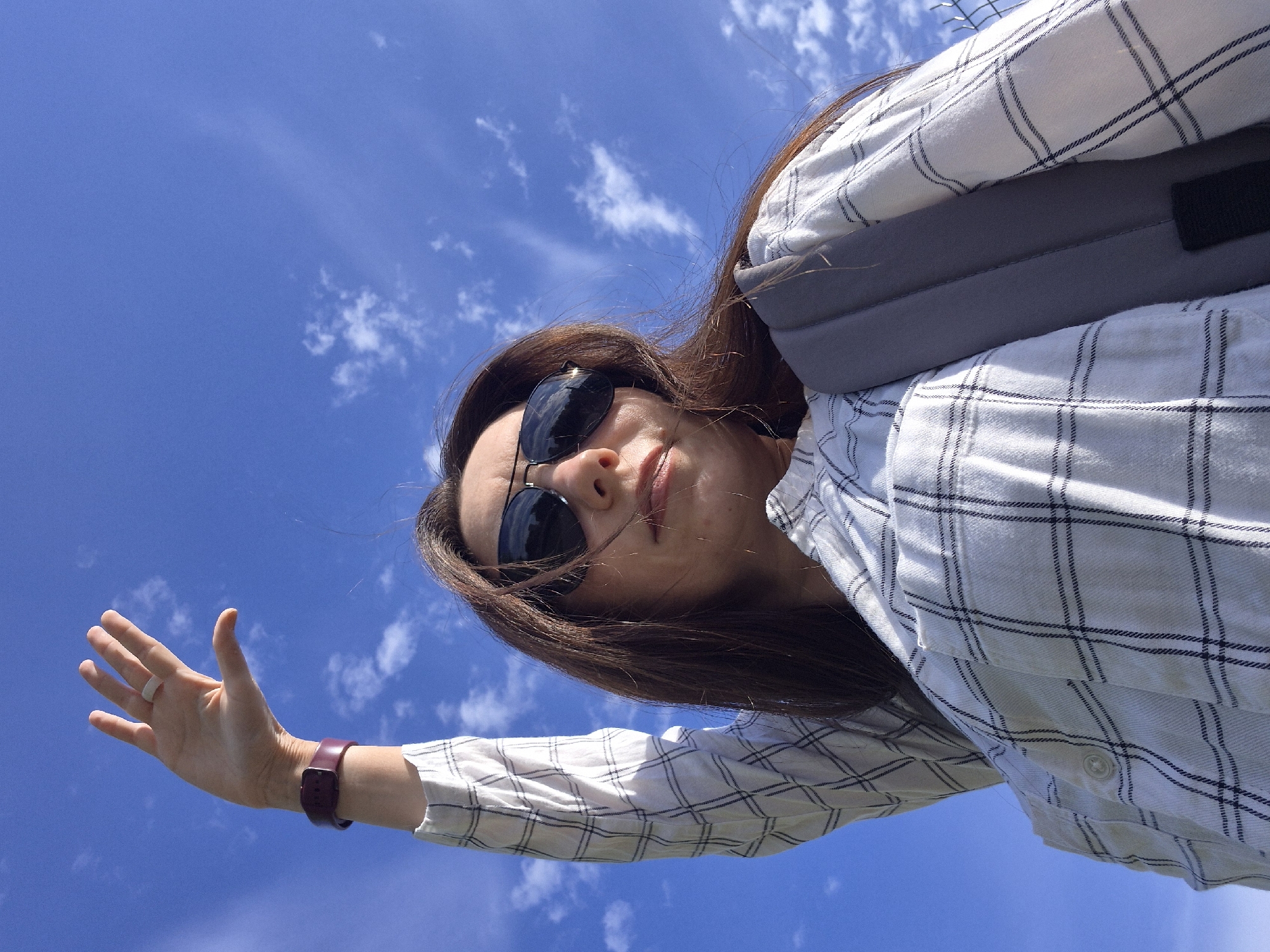 A woman stands with her face angled towards the camera, sunglasses on, against a backdrop of a blue sky with scattered white clouds. Her right arm is outstretched, hand open, and a watch is visible on her wrist. She wears a white shirt with a blue plaid pattern and has brown hair. A grey strap crosses her body, possibly from a bag. The location is unspecified.