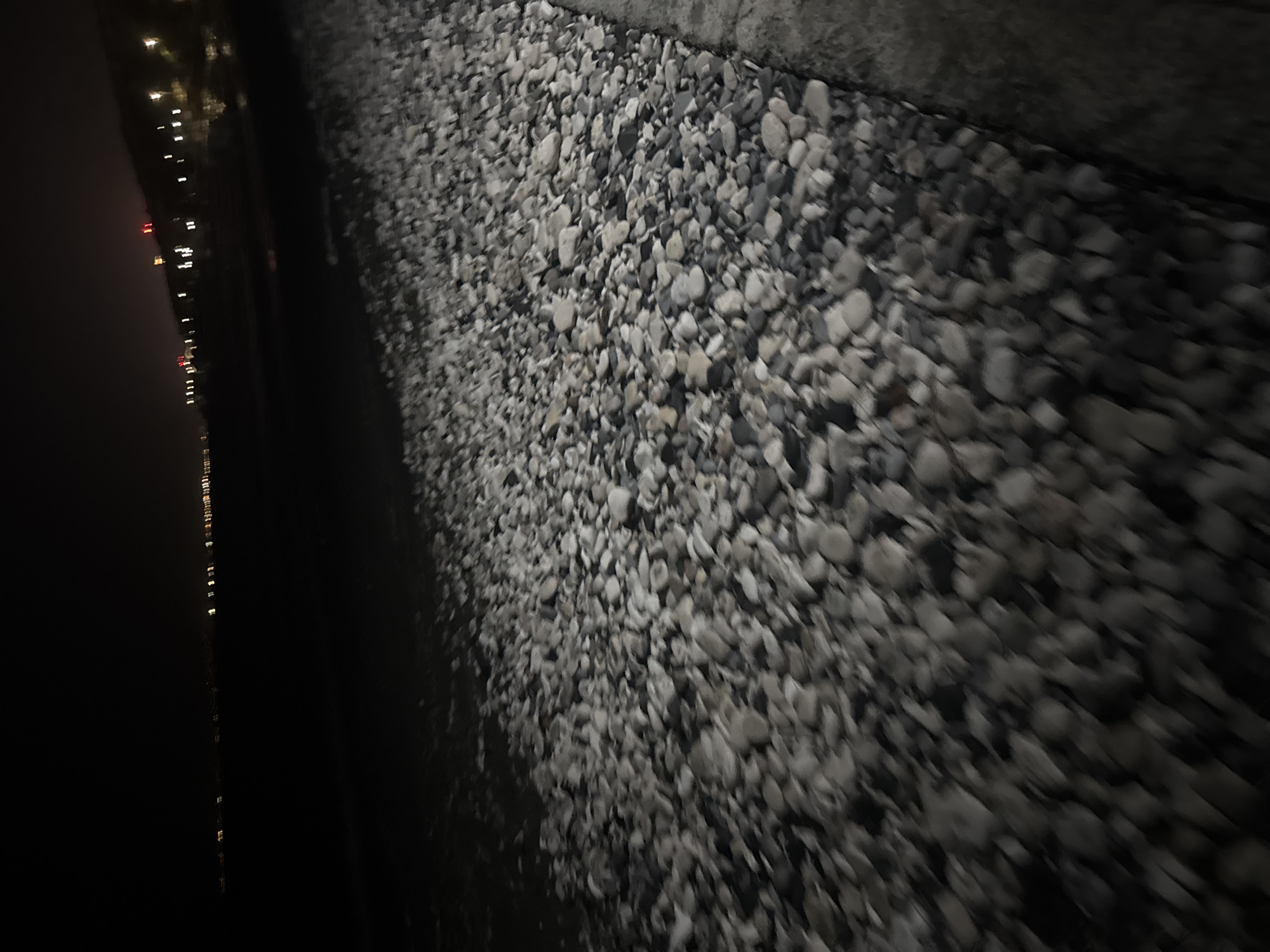 The image depicts a nighttime scene on a pebble beach in the Agios Tychona Community, Cyprus. The foreground is dominated by a dense collection of smooth, light-colored pebbles, some appearing to be wet and reflecting light. To the right, a concrete or stone walkway runs alongside the beach. In the background, the dark expanse of the sea is visible, with a faint line of distant city lights illuminating the horizon. The lights are blurred due to the darkness and distance, suggesting a coastal urban area. The overall impression is of a quiet, nocturnal beach environment with the presence of human habitation in the distance. There are no people or specific actions captured in the frame.