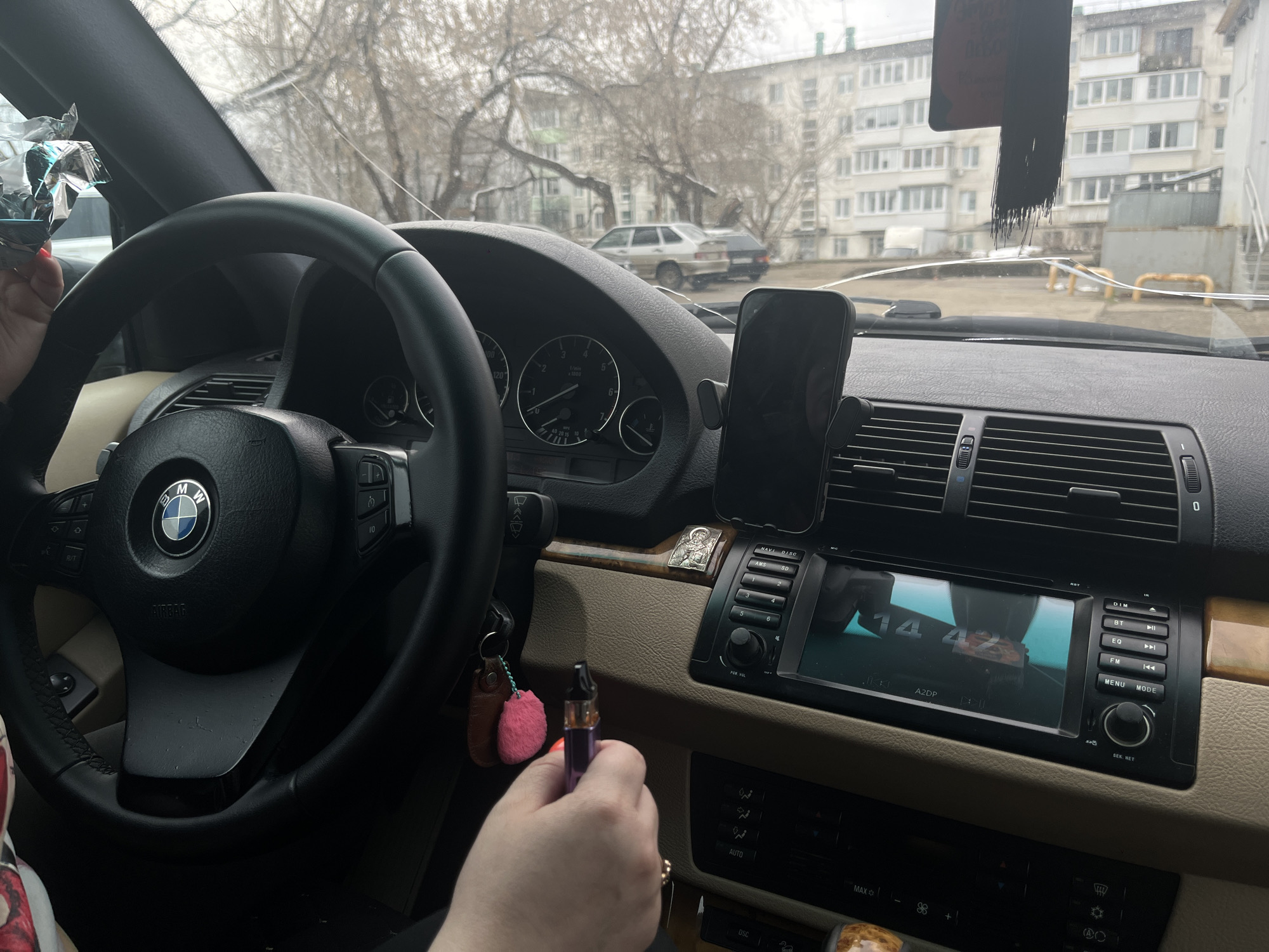 The image shows the interior of a BMW car. The driver is holding a vaping device in their right hand and what appears to be a food package in their left. A phone is mounted on the dashboard. The car is parked in what seems to be a residential area in Chaikovsky, Russia, based on the architecture visible through the windshield. The time displayed on the car's screen is 14:42.