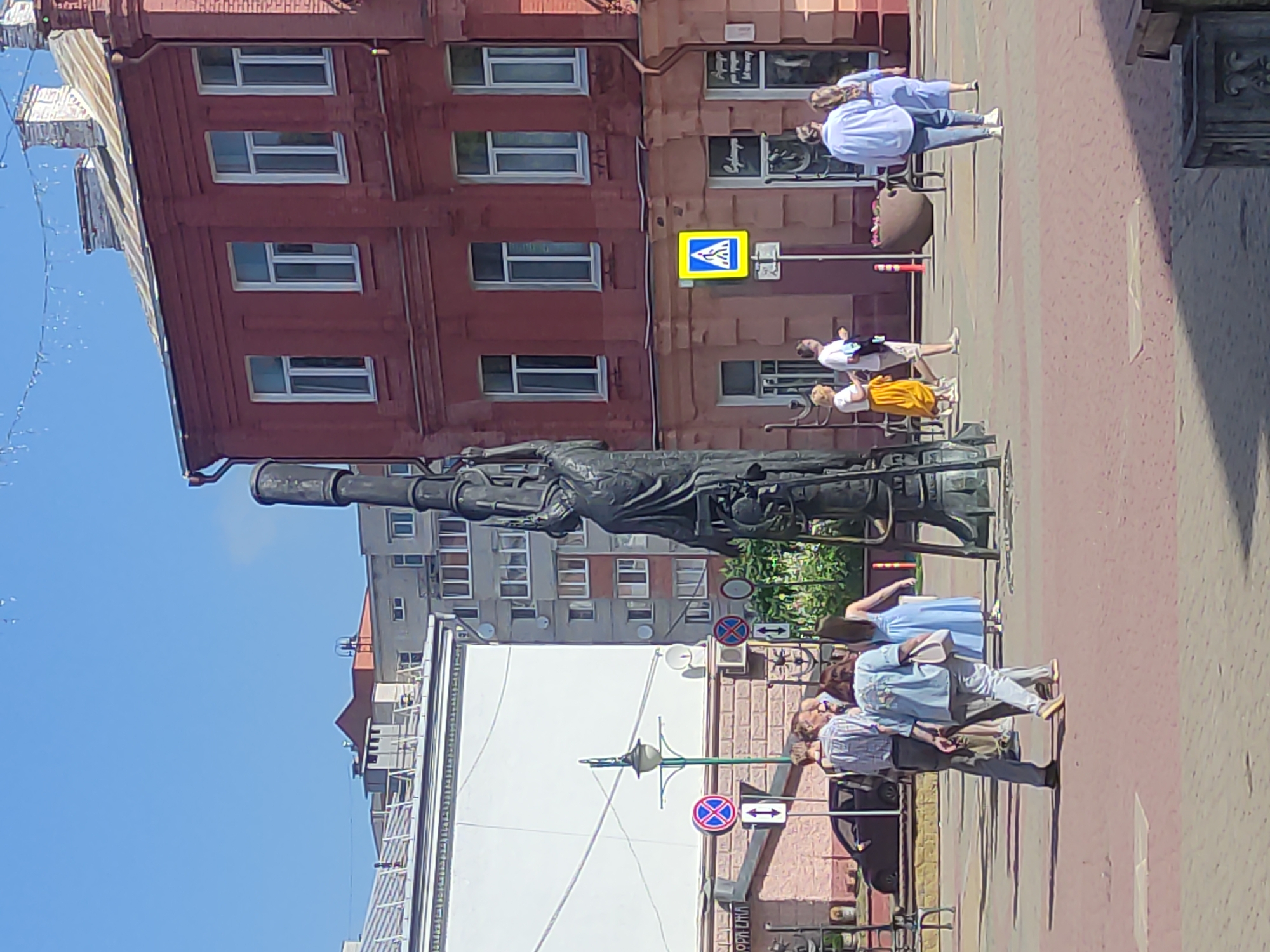 The image shows a public square in Mahilyow, Belarus, on a sunny day. A large, dark metal sculpture of a figure holding a tall, intricate structure stands prominently in the center-left of the frame. Several people are walking through the square. On the left, a white building with a large blank screen is visible, along with a street lamp and traffic signs. To the right, a red brick building with multiple windows is present, with shops on the ground floor. A yellow pedestrian crossing sign is also visible on the right. The ground is paved with red and grey tiles.