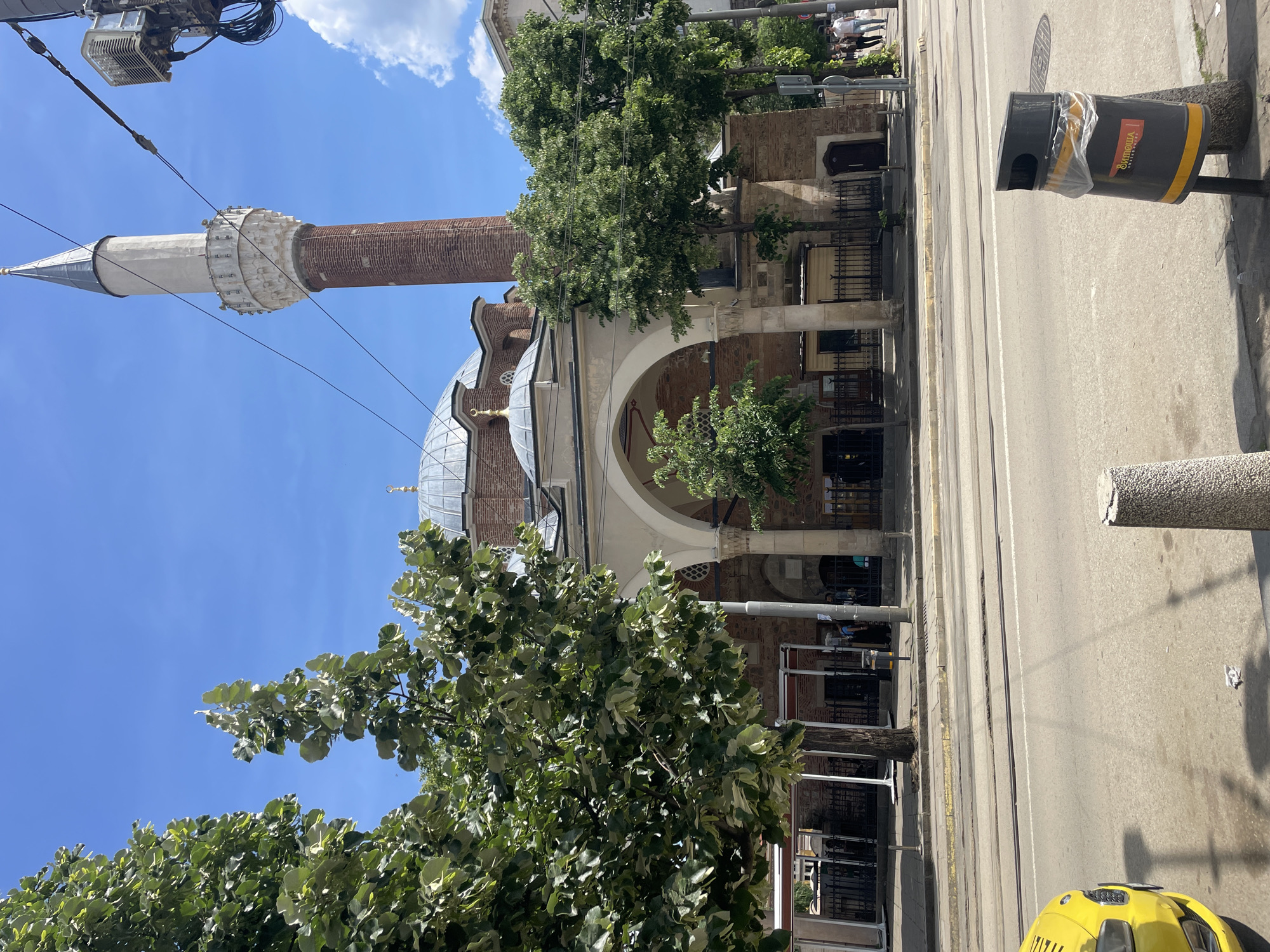 The image depicts the Banya Bashi Mosque in Sofia, Bulgaria. The mosque features a tall, brick minaret with a white, conical top, a dome, and an arched portico with columns. Trees partially obscure the view of the building. The mosque is adjacent to a street with tram lines and pedestrian traffic. A yellow taxi cab and a trash bin are visible on the street. The sky is blue with some clouds.