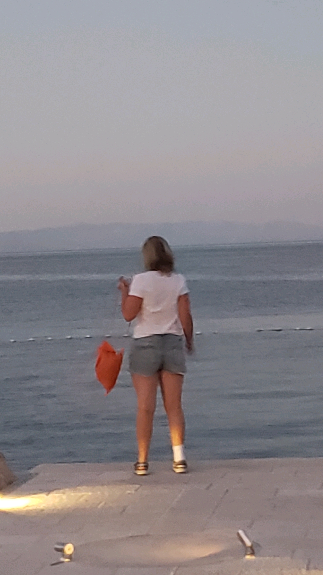 A person stands on a paved walkway overlooking the sea. The person is facing away from the camera, wearing a white t-shirt, denim shorts, white socks, and dark shoes. They are holding a bright orange bag in their right hand. The sea is visible in the background, with a faint horizon line and mountains in the distance. The walkway has a few lights and the ground is textured concrete. The scene appears to be a coastal area in Općina Tučepi, Croatia, likely during the evening or early morning given the soft light.