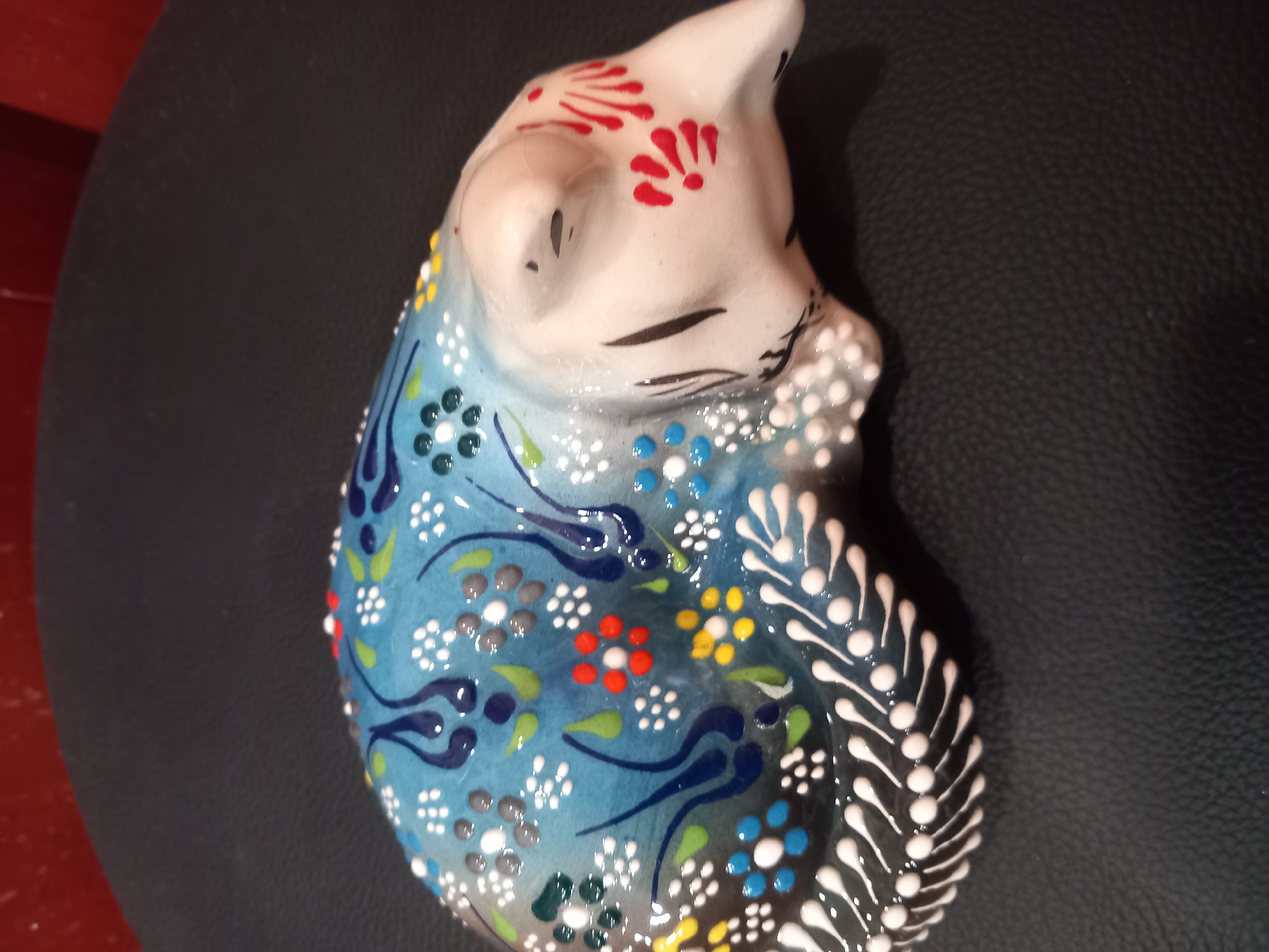 This media file shows a ceramic figurine of a cat. The cat is depicted in a curled-up position, with its eyes closed and a serene expression. Its body is adorned with intricate blue and white floral patterns, with accents of red, yellow, and green. The cat's face features red markings on its forehead, resembling petals. The figurine rests on a dark, textured surface, possibly leather or a similar material. The lighting is soft, suggesting an indoor setting. There is no discernible text in the image. The overall impression is one of a decorative art piece, possibly handmade and culturally inspired due to the detailed patterns.