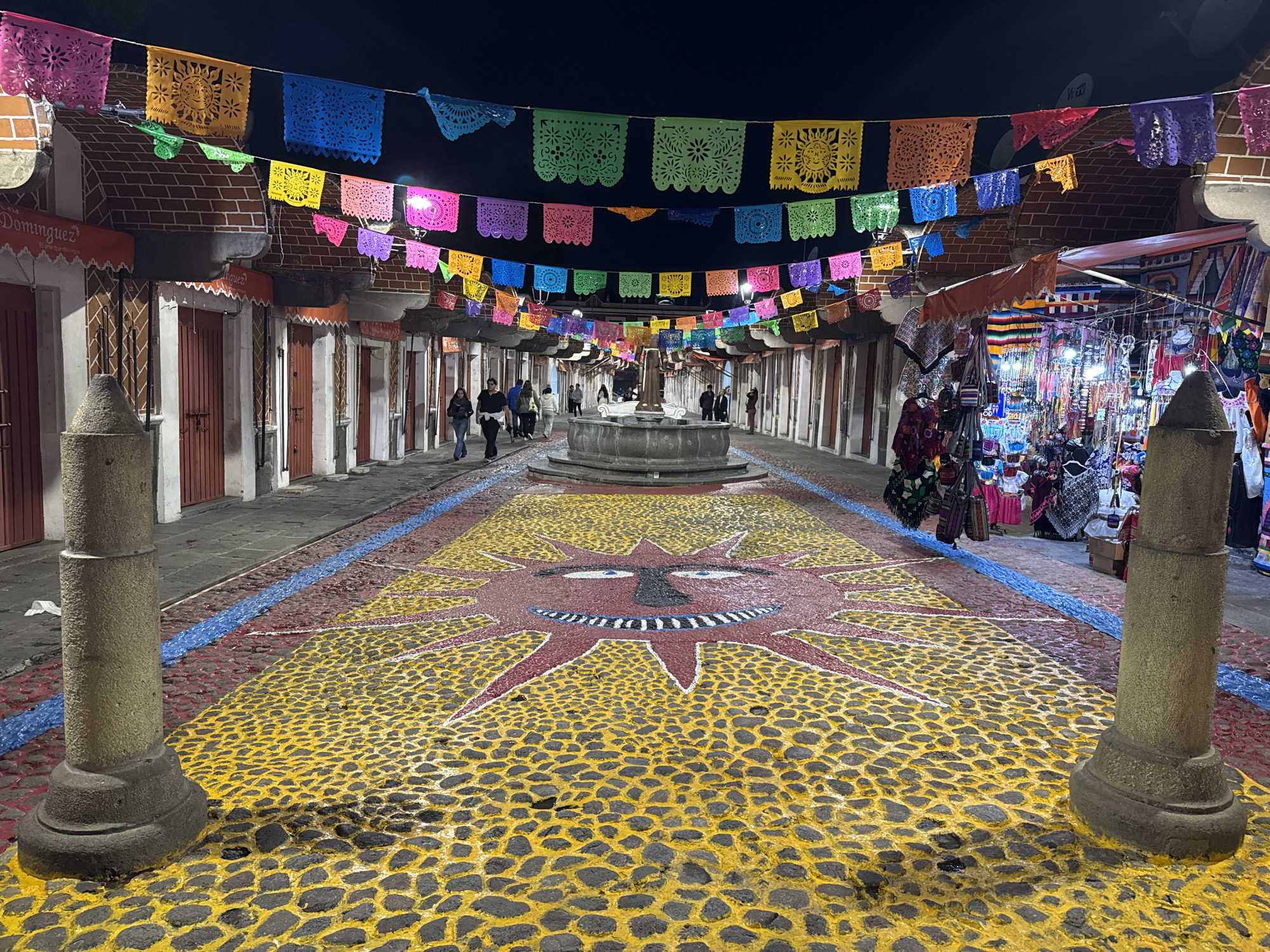 The image depicts an outdoor market street scene at night in the city of Puebla, Mexico. Rows of colorful Papel Picado banners are strung across the street overhead. On either side of the street are brick buildings with red doors, likely market stalls or shops. A stone fountain stands at the center of the street. The cobblestone street is painted with a large sun design in yellow and red. People are walking along the street. Two stone pillars frame the foreground. On the right side, a stall selling souvenirs is visible.