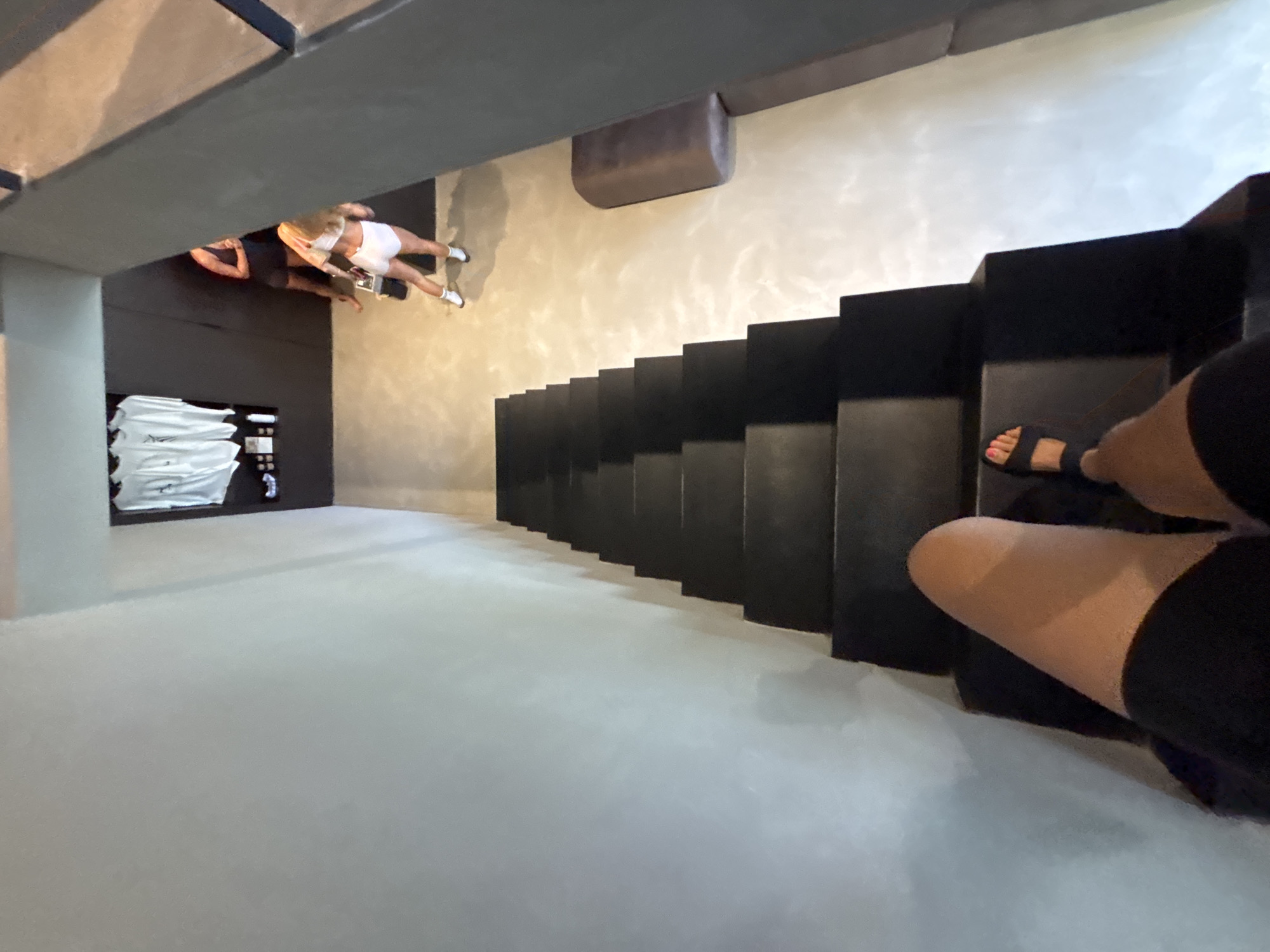 The image shows a downward-angled view of an interior space. A black staircase dominates the right side of the frame, with a person's legs and feet visible at the base of the stairs. The person is wearing black shorts and sandals.
To the left of the stairs, there's a textured wall, and in the far left corner, a display area is visible with what appears to be folded white textiles on shelves. Two people are further down the room near the left wall. One person appears to be wearing white athletic shorts and a top and the other wearing dark attire.
Given the location of Mengwi, Indonesia, and without additional information, it's impossible to determine the specific function or use of this space.