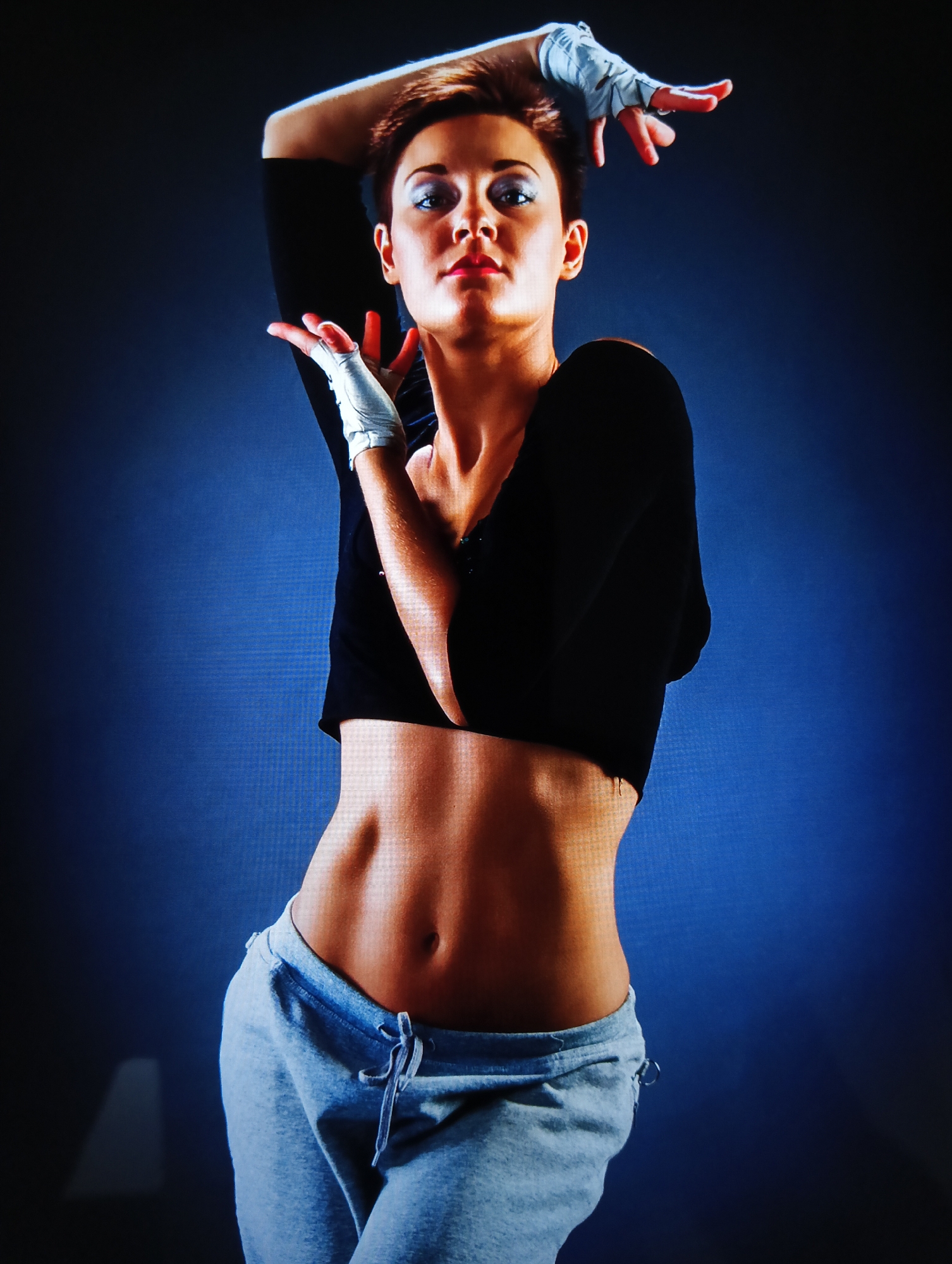 A woman is centered in the frame against a dark blue backdrop. She has a slim, toned torso, exposed by her short black crop top. She is wearing light grey sweatpants that sit low on her hips. Her hands are above her head, angled with her fingers pointing slightly outward. She has short, brown hair and what appears to be a wrap on her hands. The image does not contain enough information to determine the location.