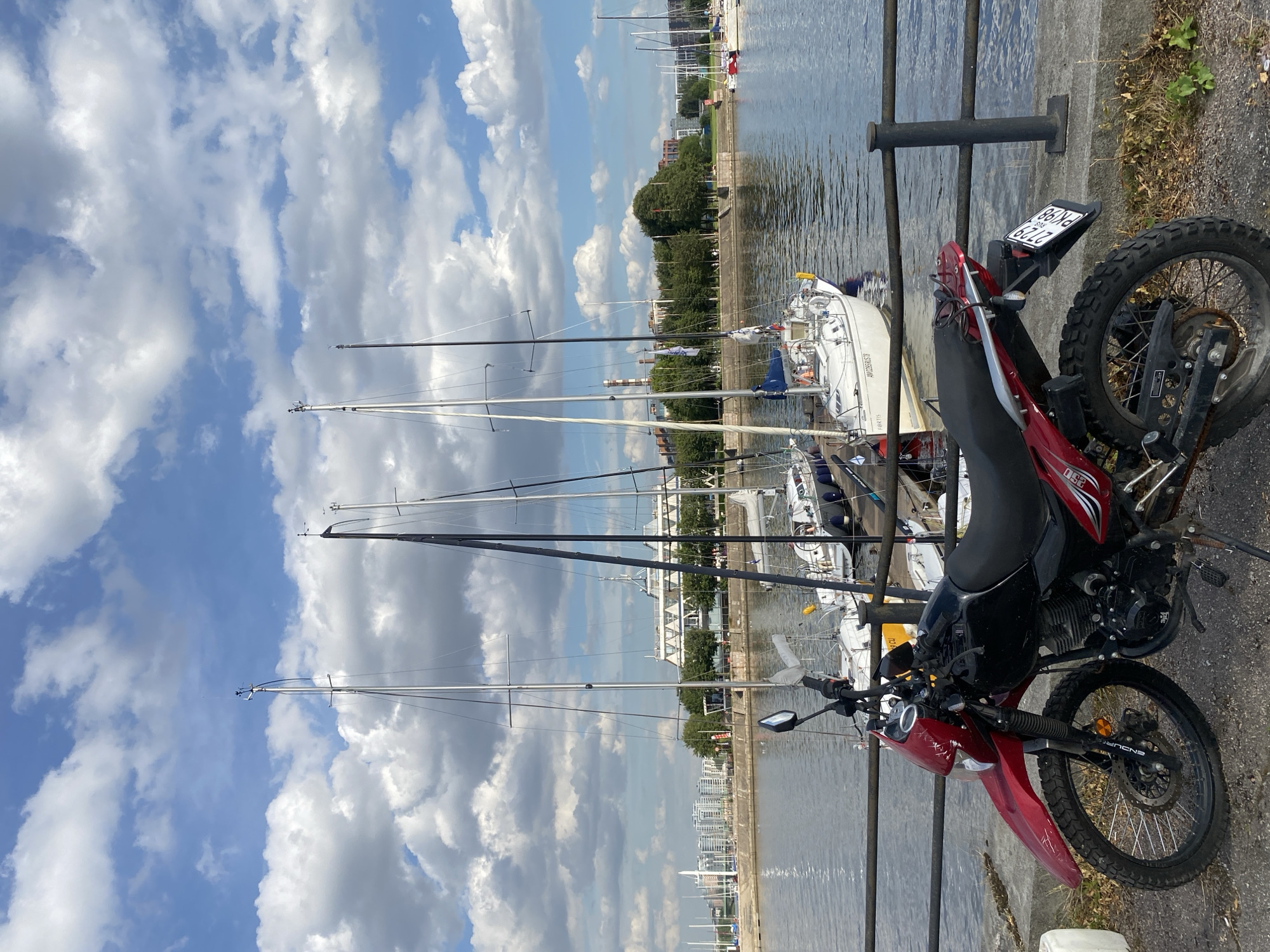 A red and black motorcycle is parked on a concrete embankment overlooking a body of water with several sailboats docked. In the background, a cityscape with buildings and trees is visible. The sky is blue with scattered white clouds. The scene appears to be in Saint Petersburg, Russia, based on the architecture and the visible license plate on the motorcycle which has Russian characters. The motorcycle is the primary subject in the foreground, positioned diagonally from left to right, with its front wheel pointing towards the left. The sailboats are moored in the water, and their masts extend upwards towards the sky. A black metal railing runs along the edge of the embankment, separating the motorcycle from the water. The overall impression is a peaceful, sunny day in a port city.