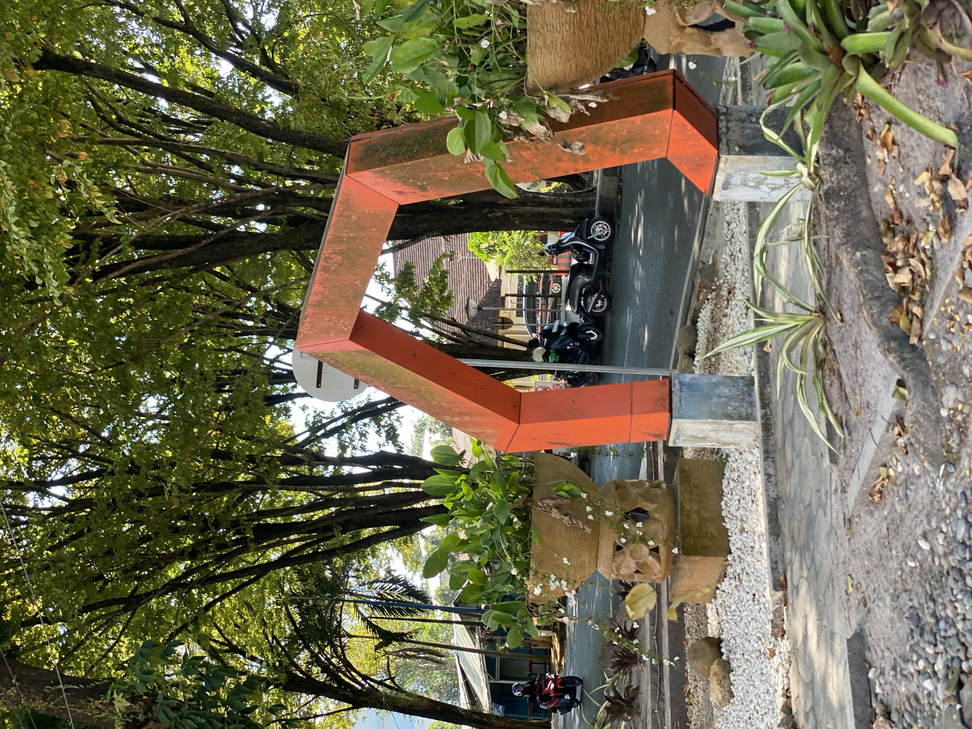 In Banjarbaru, Indonesia, an orange, angular archway stands prominently in a park-like setting. The archway is supported by concrete pillars, one of which is partially visible in the foreground. To the left of the arch, a large, decorative stone planter holds lush green plants and small white flowers, cascading down its sides. The planter is elevated on a carved stone base.
Behind the planter, a paved walkway is bordered by small white pebbles. In the background, a street is visible with several parked scooters and motorcycles. A person on a red motorcycle is seen riding on the street. Further back, there are buildings with tiled roofs and trees with dense green foliage. The environment appears to be a public area, possibly a park or a plaza, with a mix of natural elements and man-made structures. The lighting suggests it is daytime, with sunlight filtering through the trees, creating dappled shadows.