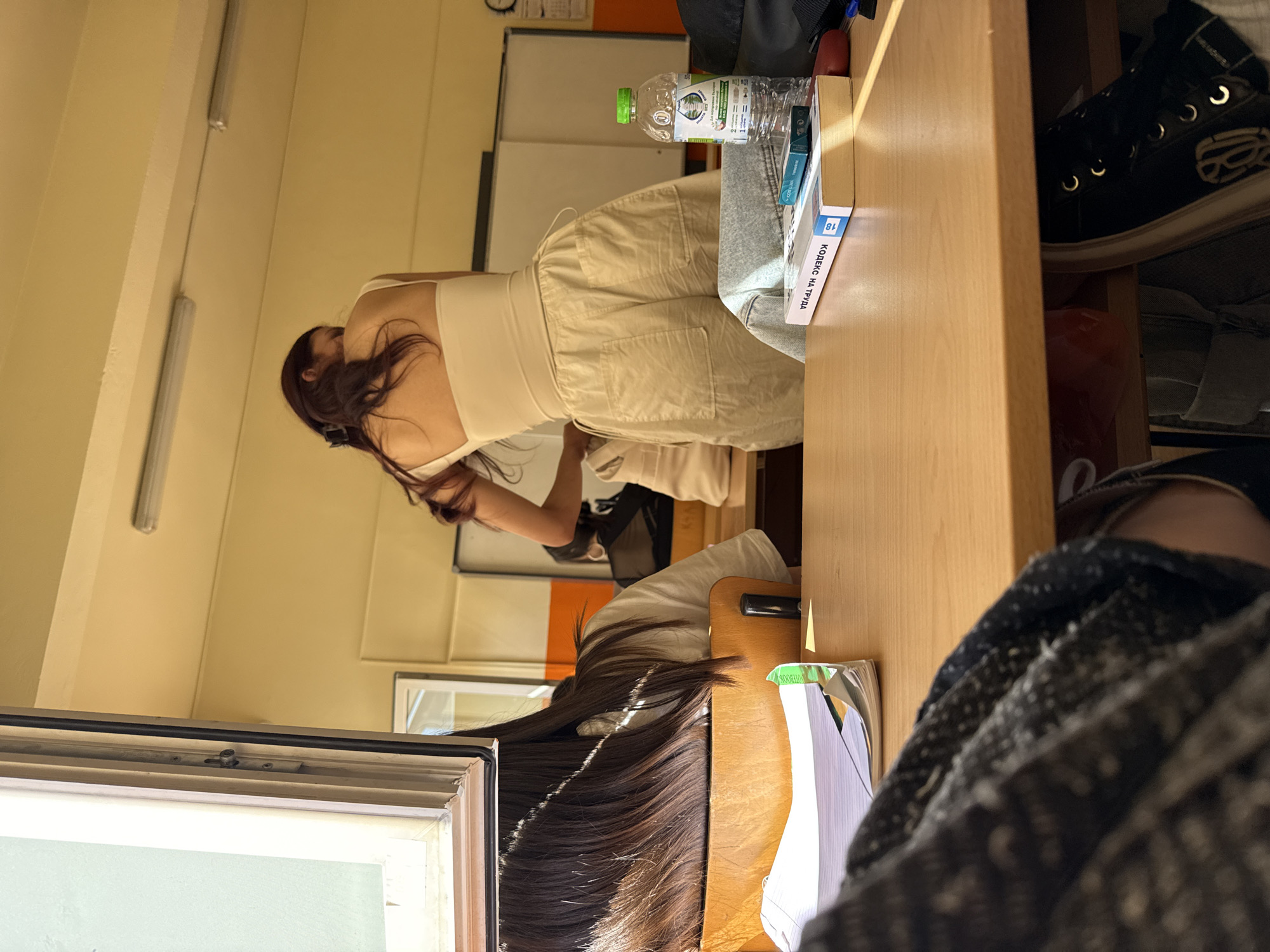 A person is bending over a desk in a room in Sofia, Bulgaria. The person has long, dark hair and is wearing a light-colored top and pants. There is a water bottle, a book, and a pair of black sneakers on the desk. The room has white walls and a window. There is a whiteboard in the background. The image is taken at an angle and is slightly blurry.