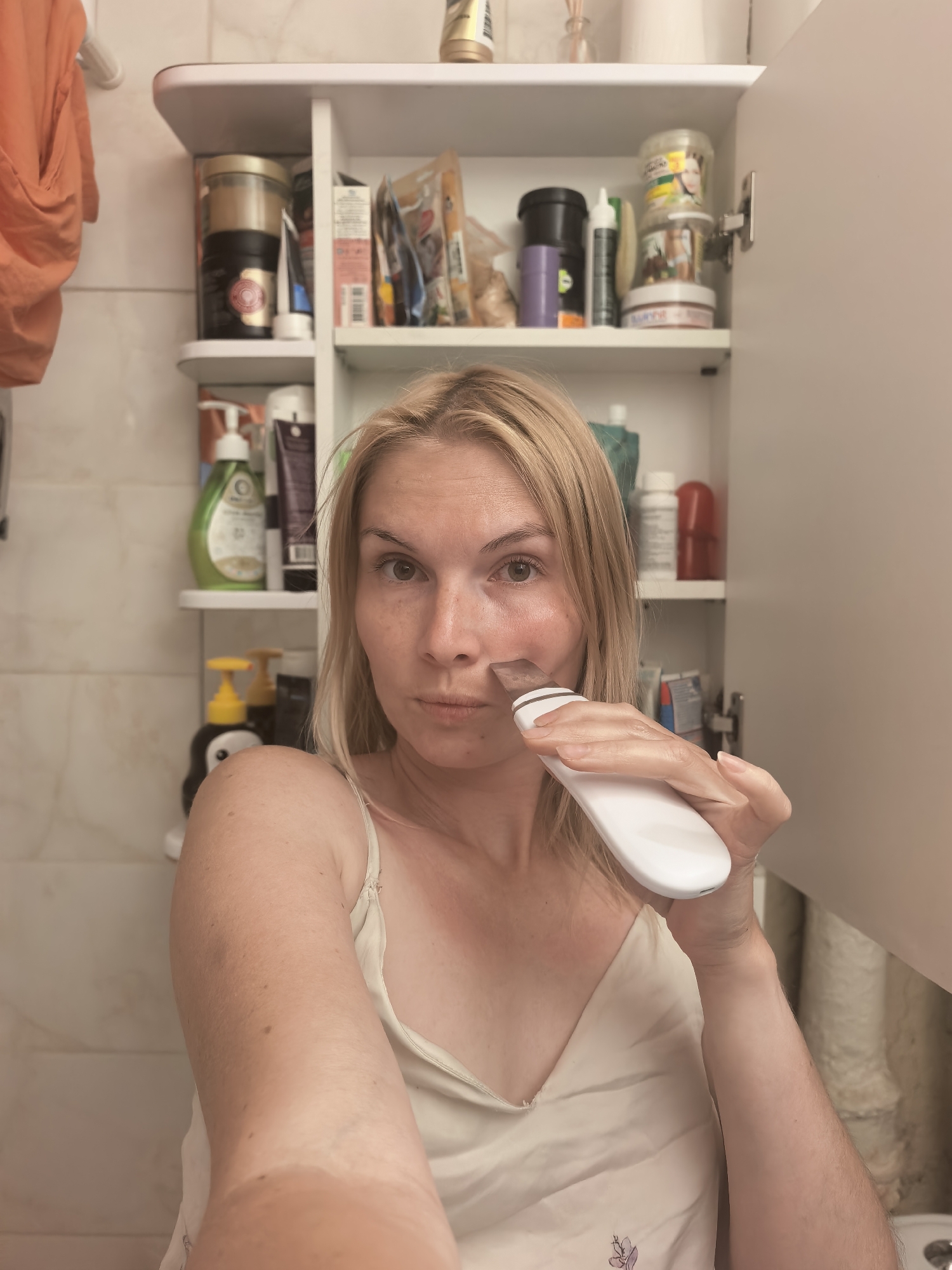 The image depicts a woman in a bathroom, holding a white electronic facial cleansing device to her cheek. She has blonde hair and is wearing a light-colored camisole. Behind her, a white cabinet is open, revealing shelves filled with various toiletries and beauty products. The bathroom walls appear to be tiled. The scene is consistent with a personal hygiene routine in a residential bathroom. There is no information in the image to suggest a specific location within the city of Sterlitamak, Russia.