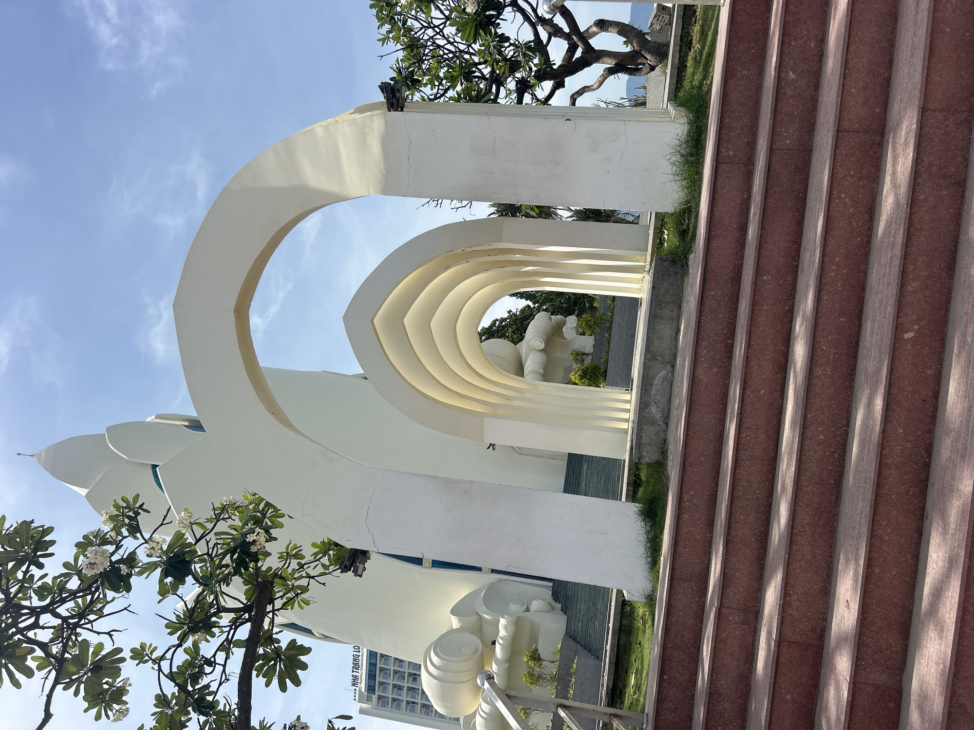 The image shows a series of white arches leading to a statue in Nha Trang Ward, Vietnam. The arches are symmetrical and decrease in size as they recede into the background. Red-toned steps are on the right side of the image. A flowering tree is visible on the left. A glimpse of the sky is visible through the arches.