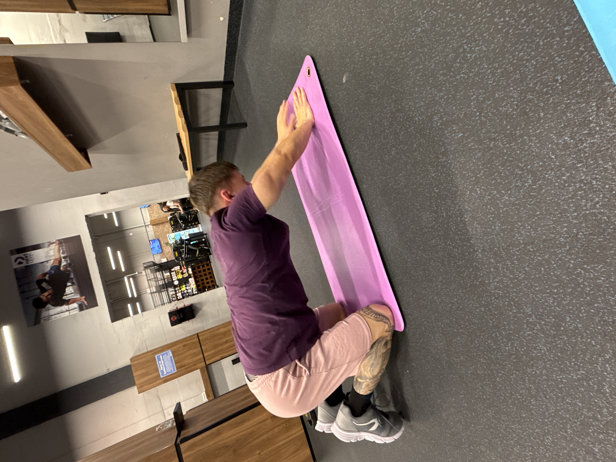 A man is kneeling in front of a purple yoga mat. He is wearing a purple shirt, pink shorts, and gray shoes. The scene takes place indoors, likely in a gym. There is a fitness center in the background, with various workout equipment and signage. The city of Sofia, Bulgaria, cannot be verified from this image.