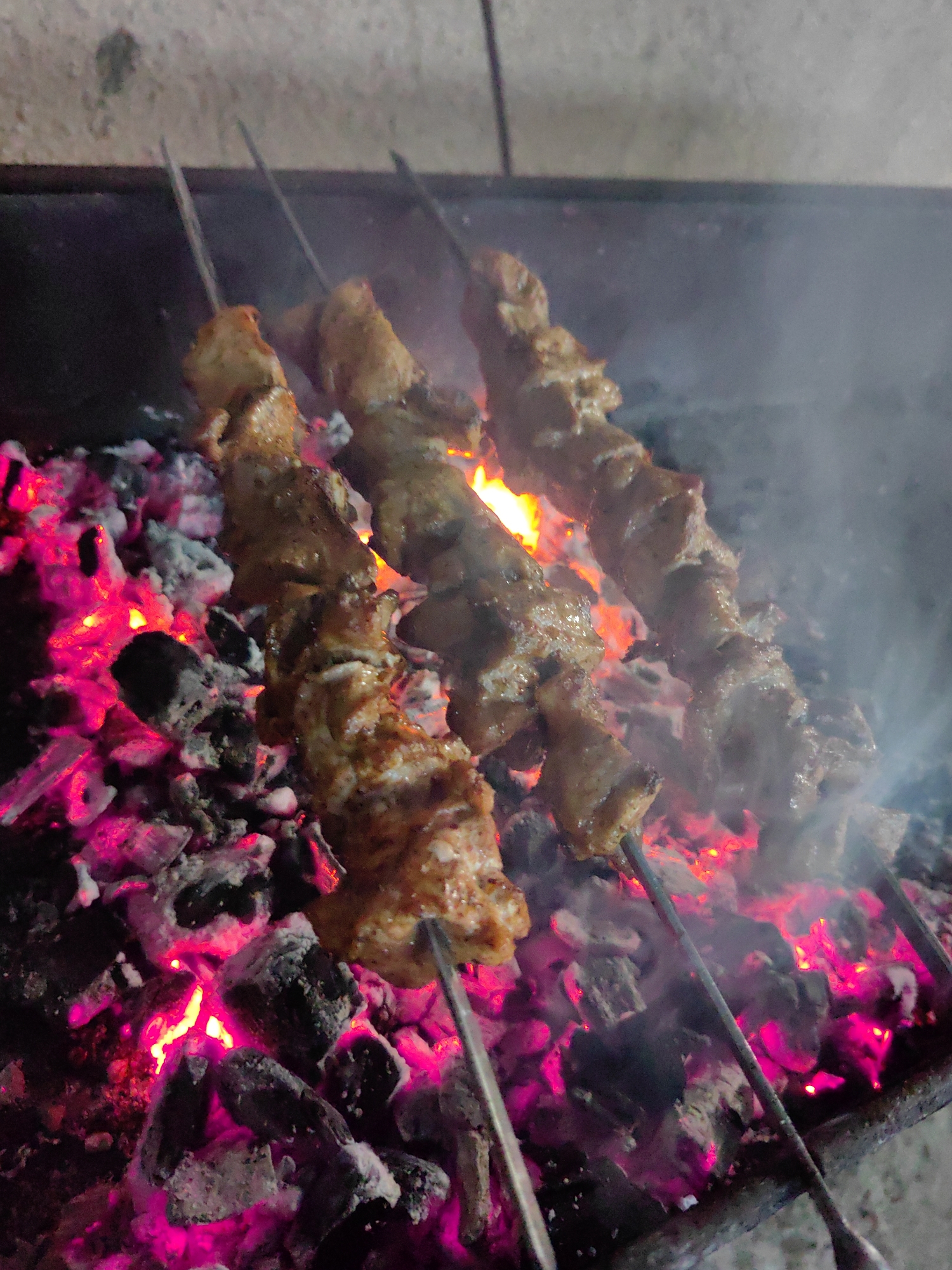The image depicts three skewers of meat being grilled over glowing pink and orange coals. The meat appears to be cubed and seasoned, with some char marks visible. Smoke rises from the coals, obscuring some of the background. The setting seems to be an outdoor barbecue or grill. There are no people or the city of Sochi mentioned or depicted in the image.