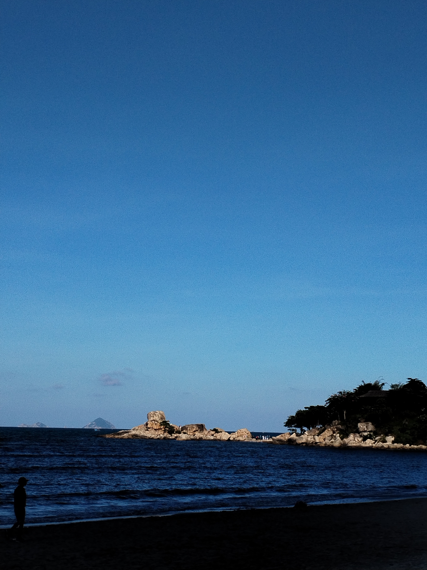 The image depicts a coastal scene, likely in or near Bac Nha Trang Ward, Vietnam. The foreground is dominated by a dark, sandy beach. The ocean extends toward the horizon, with visible waves. In the middle distance, a rocky outcrop with vegetation and what appears to be a structure stands prominently. Smaller islands are visible in the far distance. A single person is silhouetted against the ocean on the beach. The sky is clear and blue. The overall lighting suggests either early morning or late afternoon.