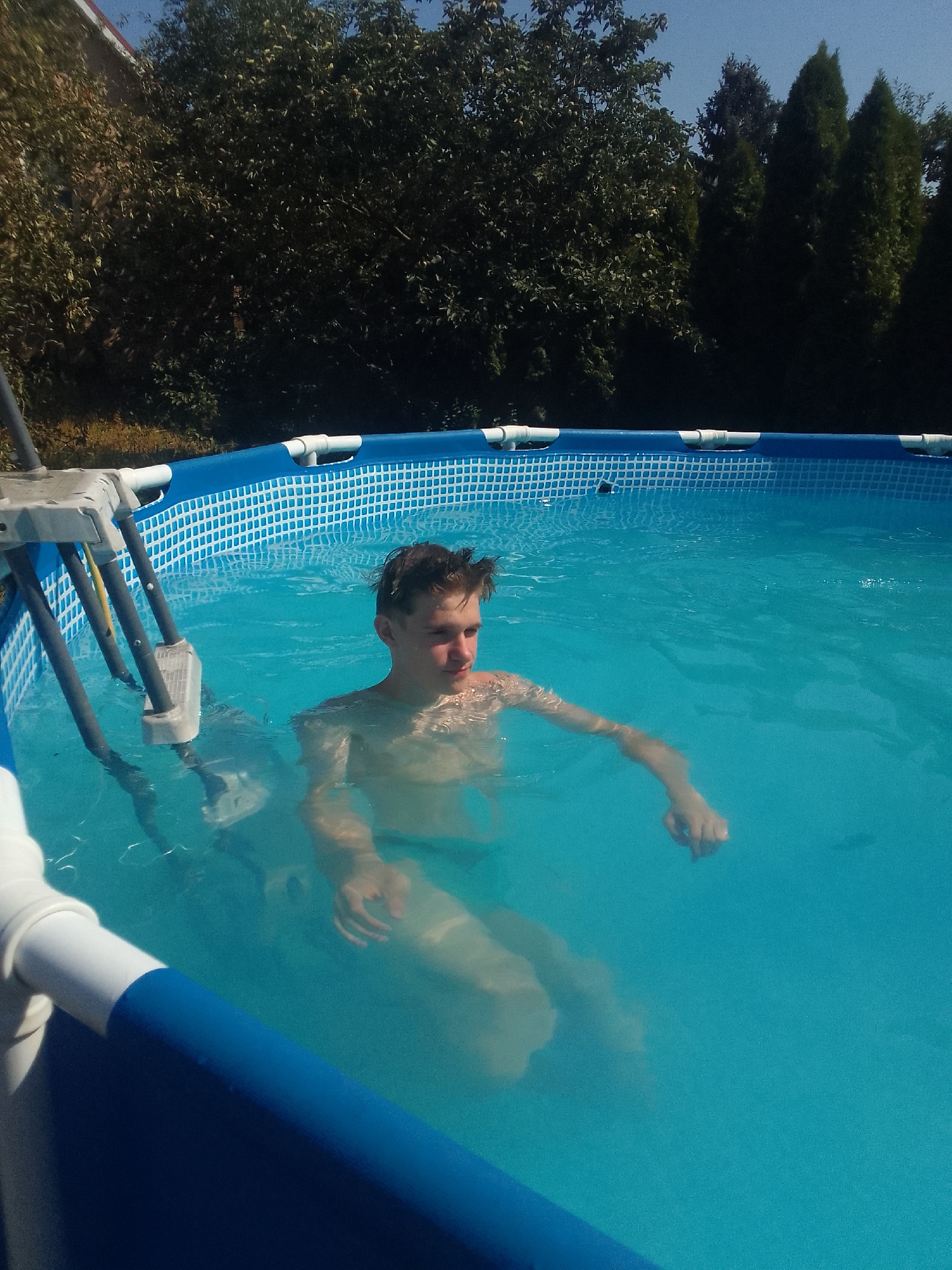 The image depicts a young male swimming in an above-ground pool filled with bright blue water. The pool is surrounded by a blue and white border, and a silver ladder with white plastic steps is visible to the left. The boy, with short, dark hair and fair skin, is submerged up to his chest and appears to be looking to his right. His arms are relaxed in the water. The background consists of lush green trees and a clear blue sky. The setting suggests a sunny day, likely in a residential backyard. This image was captured in Mynai, Ukraine.