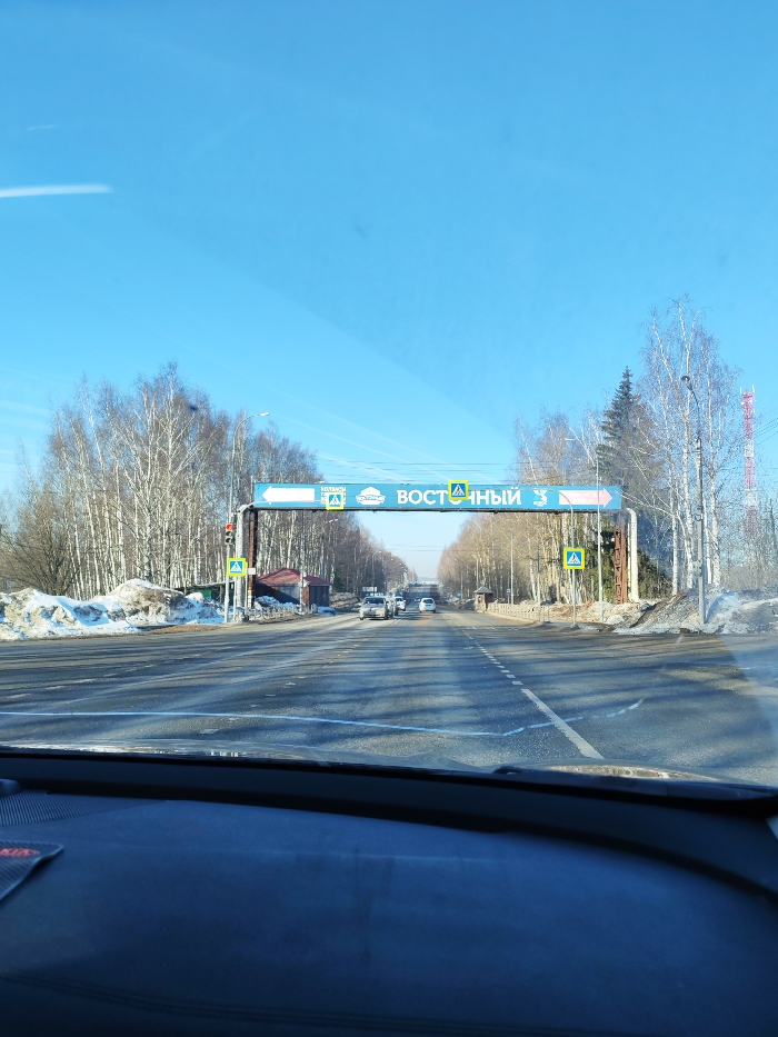 The image presents a forward-facing view from inside a vehicle on a multi-lane asphalt road in the city of Италмас, Russia, under a clear blue sky. A large blue overhead gantry sign spans the road, displaying white directional arrows and the Russian text "ВОСТОЧНЫЙ" (Eastern), along with multiple yellow pedestrian crossing symbols. Additional yellow pedestrian crossing signs are mounted on poles on both sides of the road. A red traffic light is illuminated on the left, near a gantry support. Piles of dirty snow are visible along the road shoulders. On the left, behind snow and trees, a building with a red roof is partially visible. Further down the road, several cars are observed proceeding in the same direction. The road is lined with bare deciduous trees and some evergreen trees, with a tall red and white telecommunications tower visible on the far right. The dark dashboard of the observing vehicle, including a red "KIA" emblem fragment, occupies the lower portion of the frame. No individuals are visible.