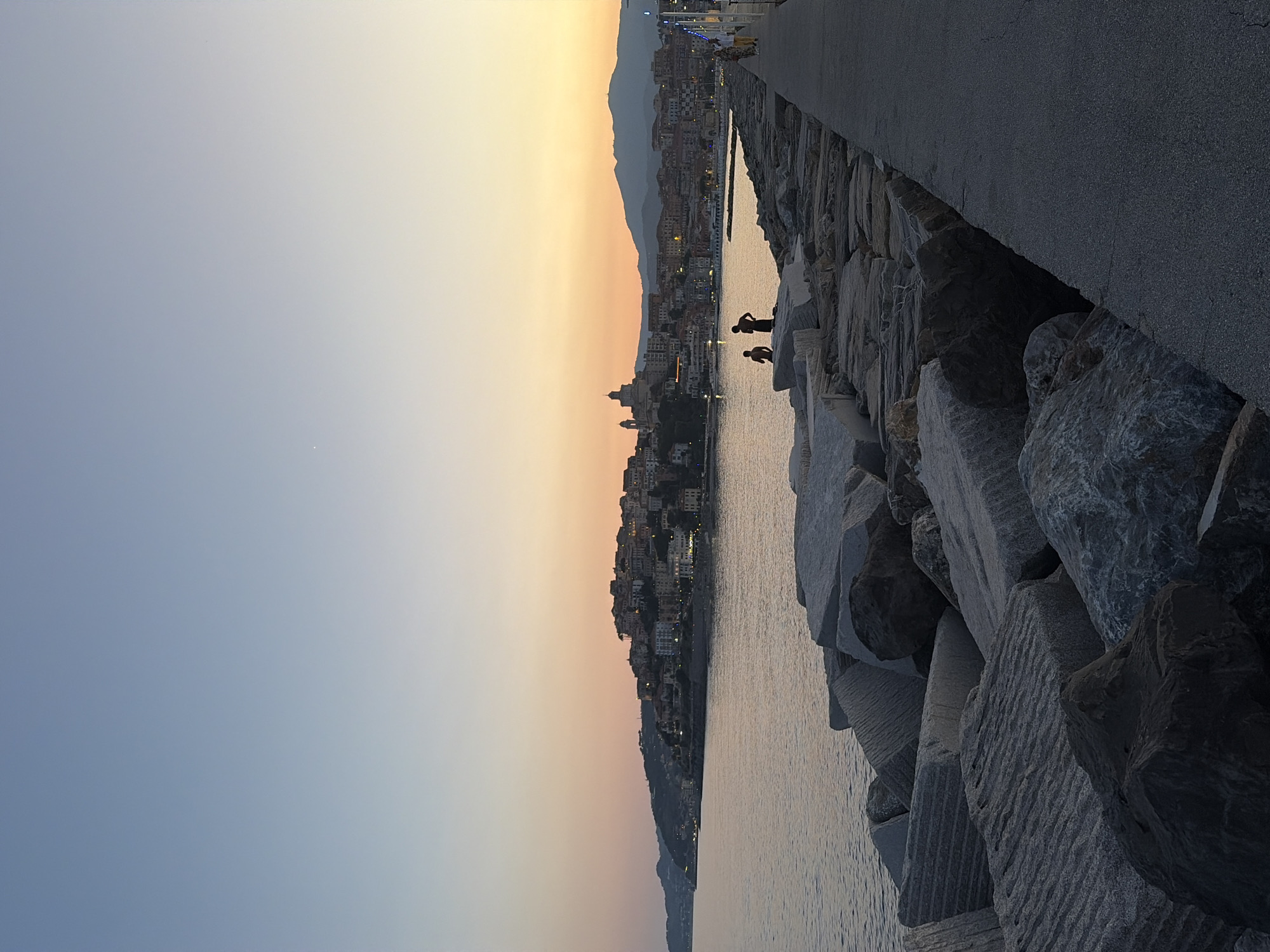 The image depicts a coastal scene at dusk in Imperia, Italy. The sky displays a gradient of colors, transitioning from a pale yellow at the horizon to a soft blue overhead, indicating sunset. The water reflects the fading light.
In the foreground, a man-made jetty or seawall is constructed of large, rough-hewn rocks. Two silhouetted figures are visible on the rocks near the water's edge, appearing to be interacting or observing the surroundings. Further along the jetty, a paved walkway runs parallel to the rocky structure.
Beyond the water, a town is nestled against a backdrop of hills. The buildings of the town are illuminated by lights, suggesting that evening has fallen. The lights of the town twinkle against the darkening landscape. The overall atmosphere appears serene and peaceful, capturing the tranquil beauty of the Italian coast at twilight. No visible text is present in the image.