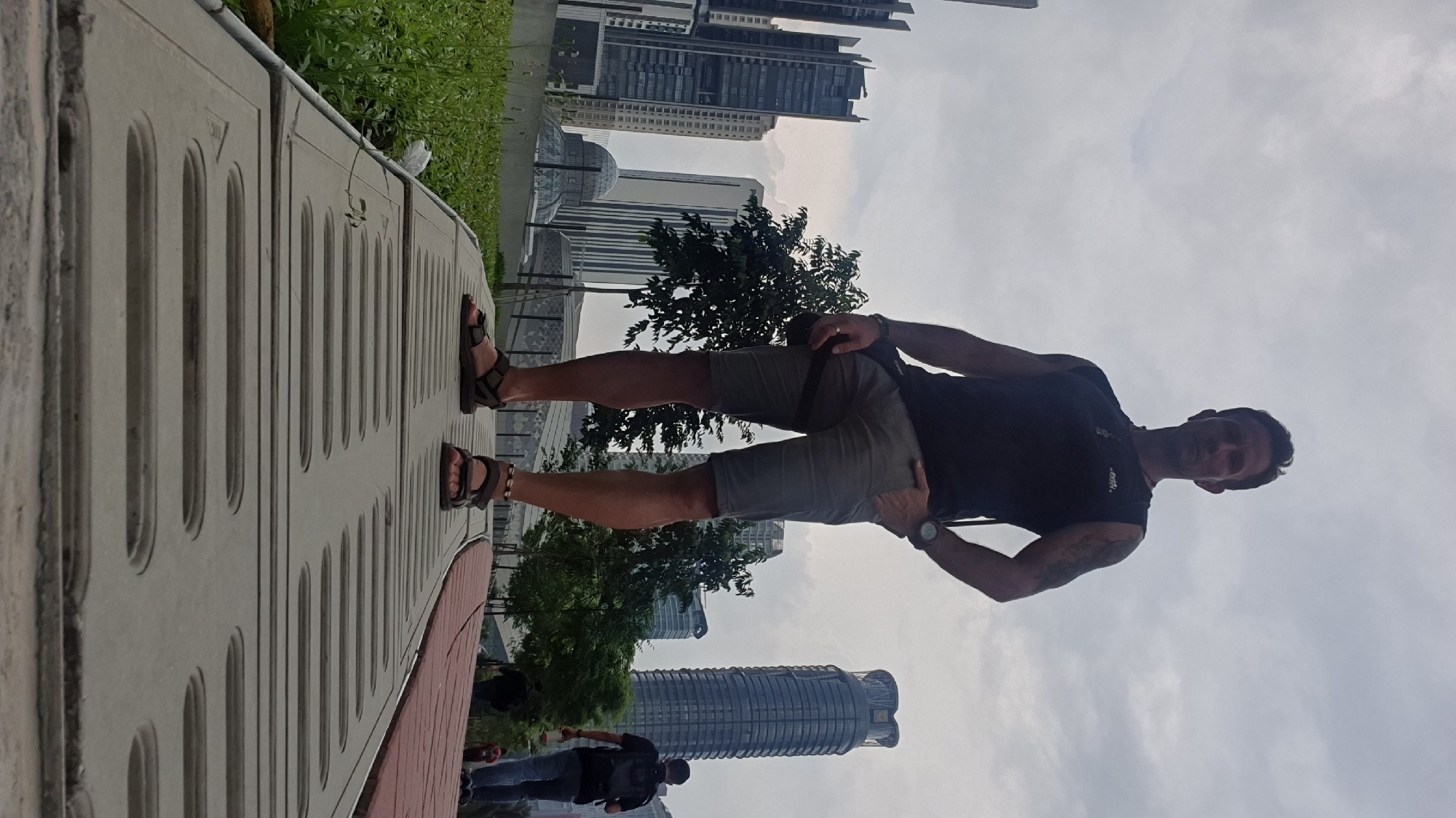 A man stands in a park in Kuala Lumpur, Malaysia, with a cityscape in the background. He is viewed from a low angle, looking up at him. He is wearing a black sleeveless shirt, grey shorts, and dark sandals. He has a watch on his left wrist and a bag slung across his chest. His right hand is in his pocket. In the background, there are several tall buildings, including a distinctive circular skyscraper, and some trees. The sky is overcast. The man appears to be posing for a photo. There are other people in the distance, walking in the park.