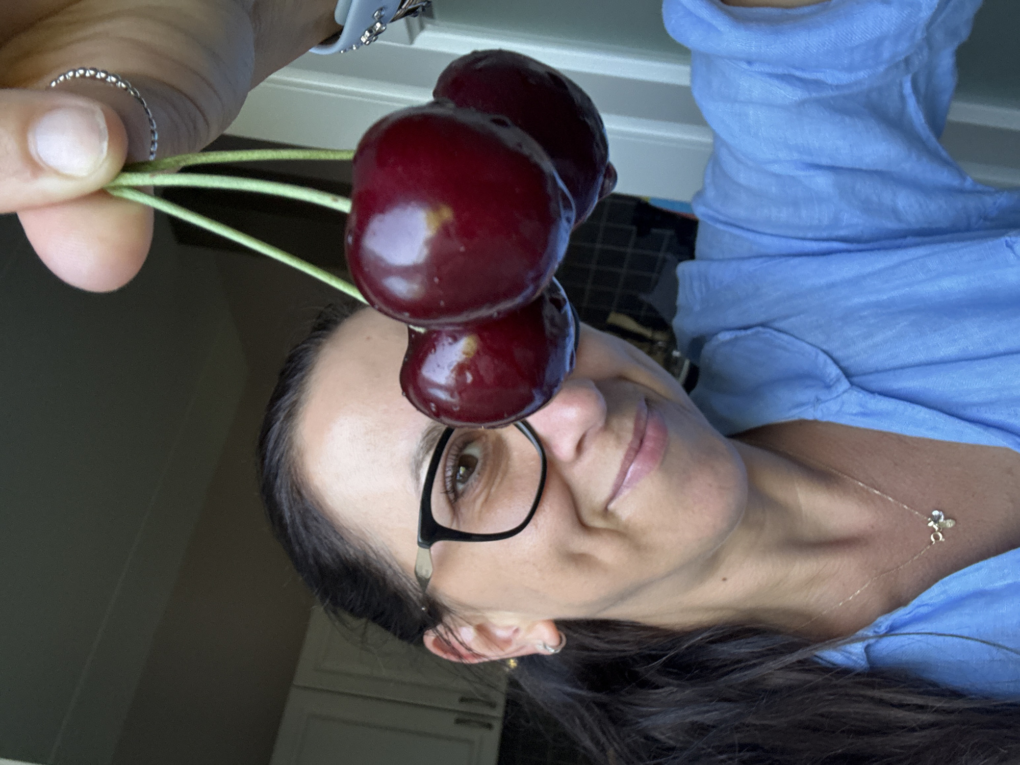 In Marum, Netherlands, a close-up, self-portrait captures a woman wearing glasses and a blue collared shirt. She is holding a cluster of three dark red cherries by their stems in her right hand, with the cherries obscuring part of her face. A delicate necklace with a small pendant is visible on her neck. The background includes kitchen cabinetry and a light-colored door.