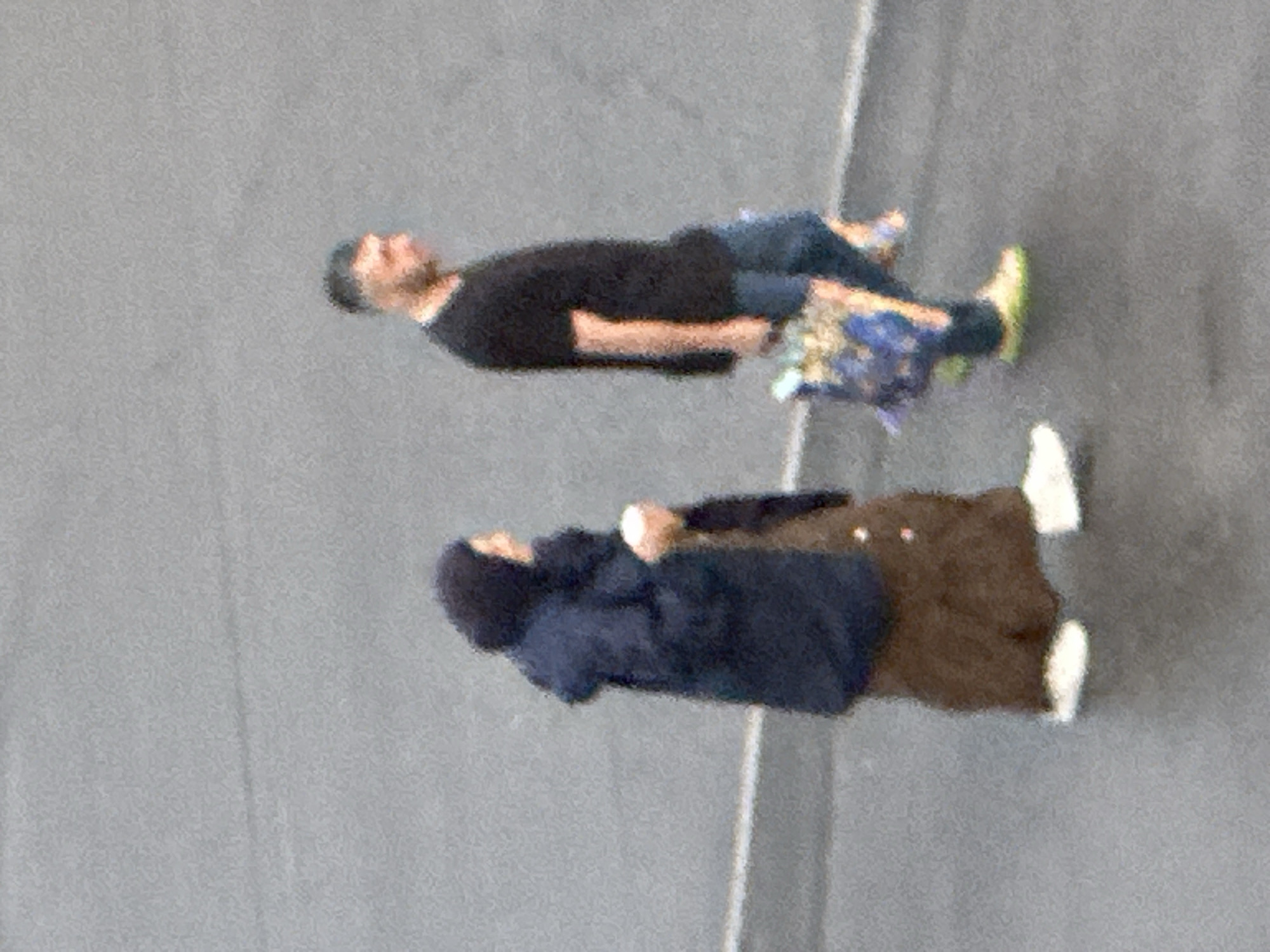 Two individuals are walking on a grey, paved surface, likely a street or sidewalk in Podolsk, Russia. To the left, a person wearing a dark hooded jacket and a brown skirt or trousers is walking away from the camera, holding a light-colored cup in their right hand. To the right, a person wearing a black t-shirt and blue jeans is walking towards the right, carrying a bag with colorful items. The pavement has a faint white line running horizontally across it. The overall lighting suggests an overcast day.