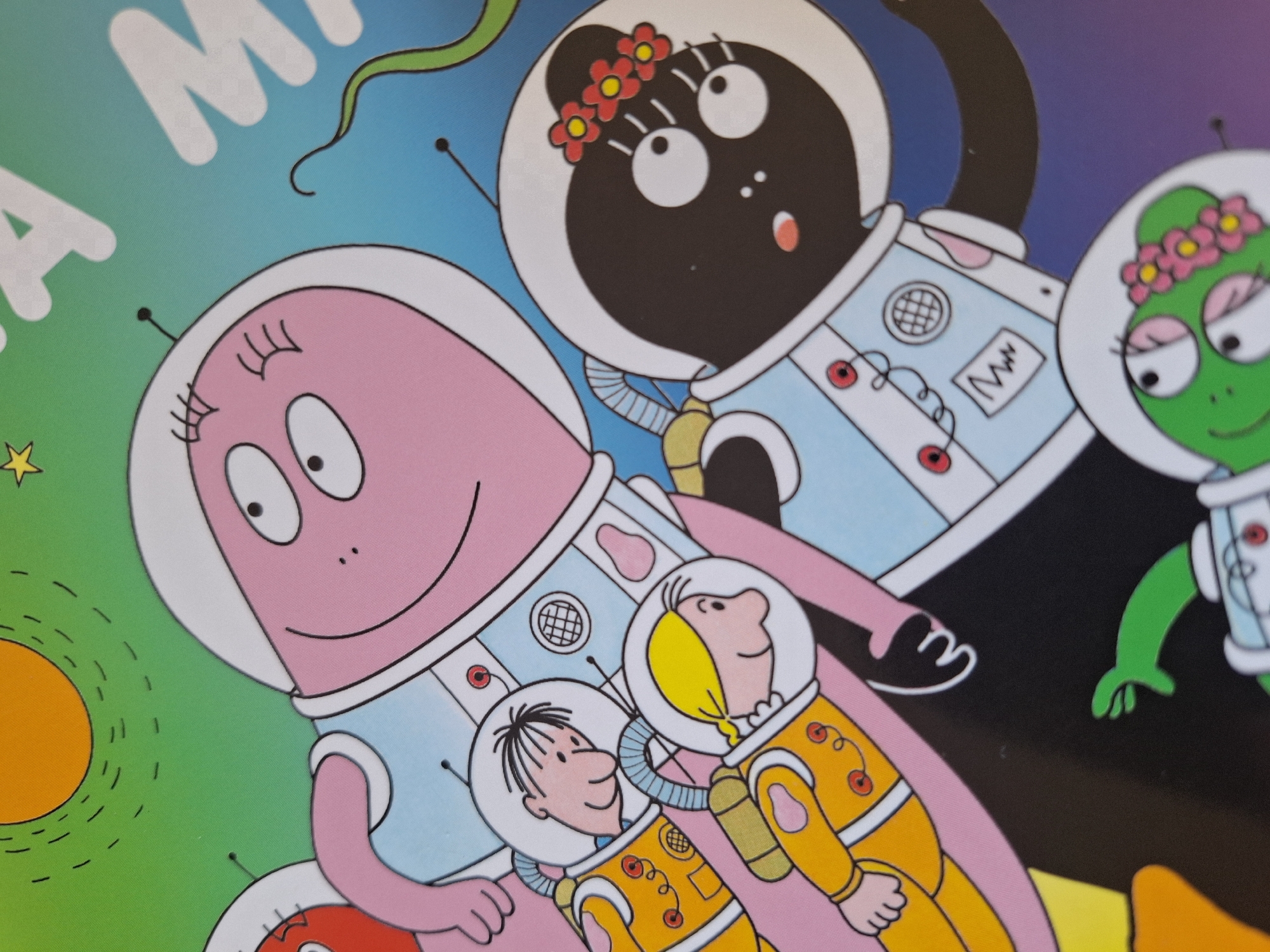 The image shows a cartoon illustration featuring characters in space suits. The background transitions from green with a yellow sun and dotted lines on the left to blue and purple on the right. A pink, oval-shaped character is positioned in the foreground wearing a light blue and white space suit. Below the pink character are two human-like figures in yellow and white space suits. Above and to the right of the pink character is a black, oval-shaped character with a flower crown, wearing a light blue and white space suit. To the right of the black character is a green, oval-shaped character, also wearing a light blue and white space suit with a flower crown. Part of a white word is visible at the top left of the image. The scene depicts an outer space theme with cartoon characters in astronaut attire. Location: Unknown, Unknown.