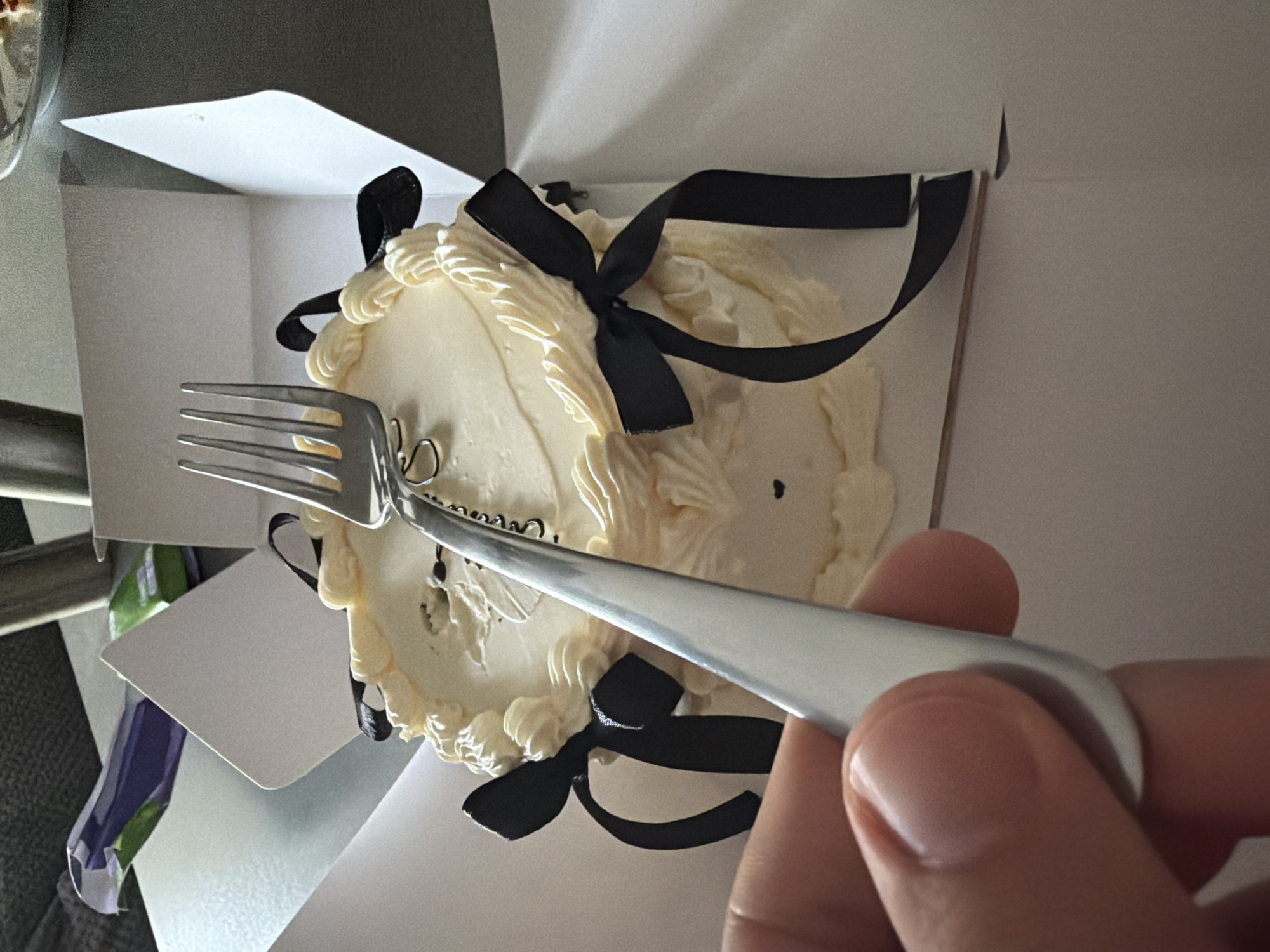 The image depicts an indoor scene focused on a small, round cake within an open white cardboard box. The precise location within the city of Sofia, Bulgaria, cannot be determined from the provided visual information.
The cake is covered in smooth, light-colored (white or off-white) frosting, with additional piped frosting borders along its top edge. Four black satin ribbons are tied into bows and attached symmetrically around the cake's perimeter. A small, dark, possibly heart-shaped detail is visible on the side frosting. The top surface of the cake exhibits a slight, irregular depression or mark.
In the lower-left foreground, a human hand with a light skin tone is visible, holding a silver-colored metal fork. The fork's tines are pointed downwards, positioned directly over the center-right portion of the cake's top surface. The hand appears poised to interact with the cake.
The background is out of focus, showing a dark gray upholstered surface in the upper-left corner and a light-colored surface, possibly a table or counter, behind the cake box. Indistinct, blurred elements, including what appears to be purple and green packaging, are also present in the background.