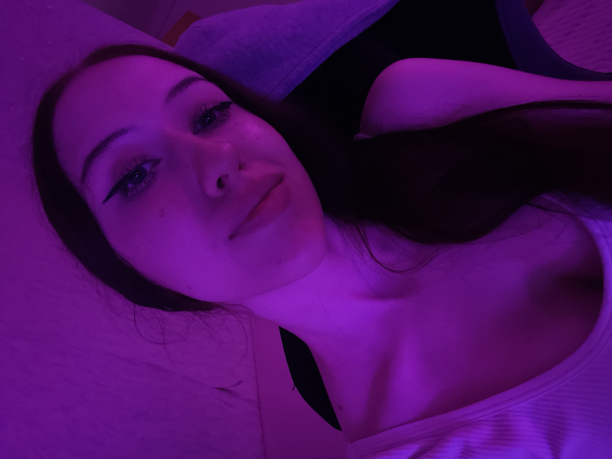 The image is a close-up, low-angle selfie taken indoors, illuminated by purple lighting. A young woman with long dark hair, styled in waves, is the sole subject. She is wearing a white ribbed tank top. Her face is tilted, and she is looking directly at the camera with winged eyeliner and full lashes. The background is indistinct, appearing as a textured surface in shades of purple, possibly a wall or fabric. The overall atmosphere is intimate and suggestive. There is no discernible information about the location being in Votkinsk, Russia.