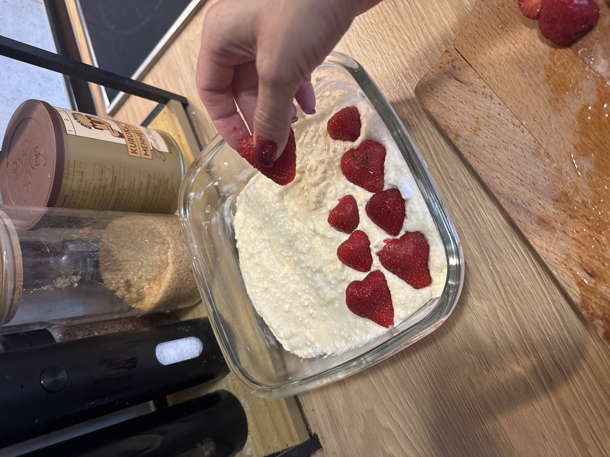 A close-up shot shows a hand with fingernails painted purple, holding a sliced strawberry above a glass baking dish filled with a creamy white dessert. The dessert is decorated with several sliced strawberries. In the background, there are glass jars filled with brown sugar and a can of Turkish coffee. The scene appears to be set on a wooden countertop.