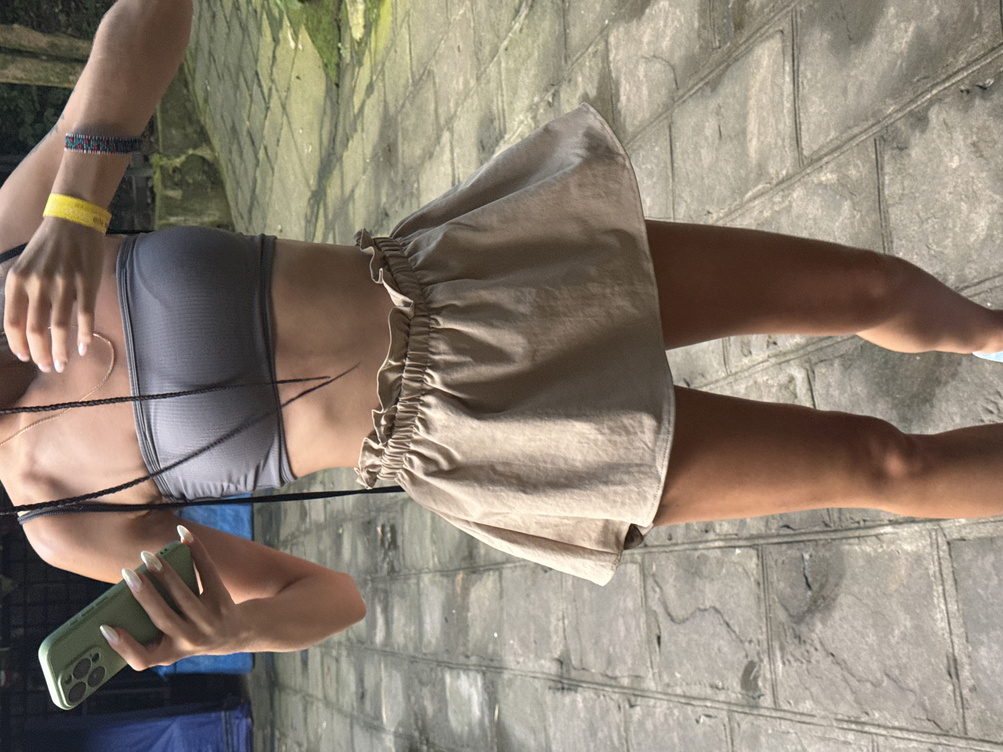A woman is standing outdoors, holding a smartphone. She is wearing a grey tube top and a beige ruffled skirt. Her hair is styled in braids. She has bracelets on her right wrist, one yellow and one multi-colored. The ground is paved with stone tiles. In the background, there is greenery and what appears to be a blue tarp. The scene is likely in a park or natural setting within Tây Hoa Lư Ward, Vietnam, given the paving stones and foliage.