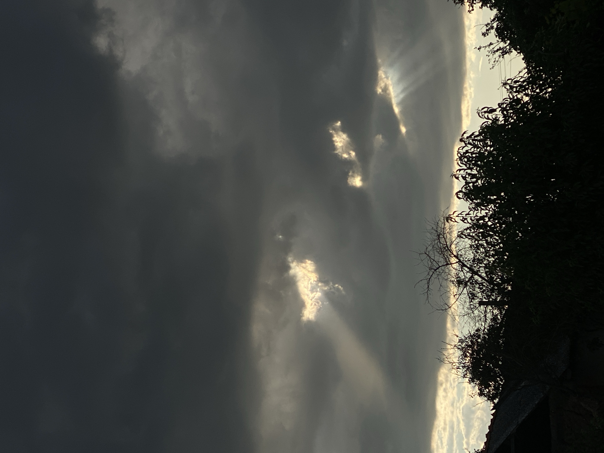 The image depicts a dark, cloudy sky with beams of sunlight breaking through in patches. The sun's rays are visible as bright shafts cutting through the dense, grey clouds, illuminating small sections of the sky with a yellowish glow. The lower portion of the image is dominated by the dark silhouettes of trees and a portion of a rooftop, suggesting an outdoor scene at dusk or during stormy weather. The overall mood is dramatic and slightly foreboding, with the light piercing through the oppressive darkness. The location is given as Cairaclia, Moldova.