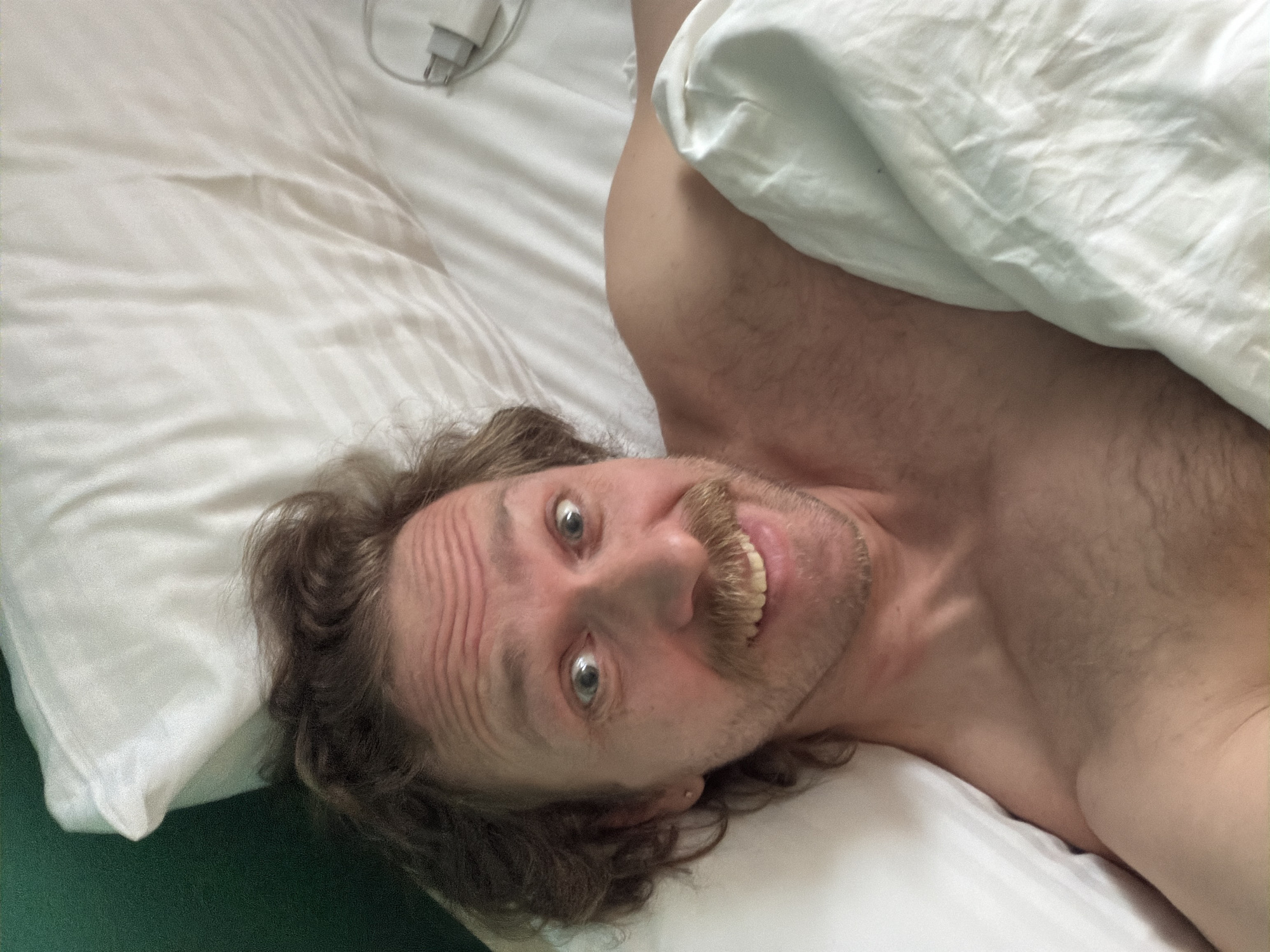 A man with shoulder-length curly brown hair and a mustache lies on a bed with white sheets and pillowcases. His head is tilted to the side, and he is looking at the camera with wide eyes and a smile. His upper torso is visible, revealing a hairy chest. A charging cord and adapter rest on the white sheet beside his head. The image was most likely taken indoors. The location cannot be determined as Minsk, Belarus, without further information.